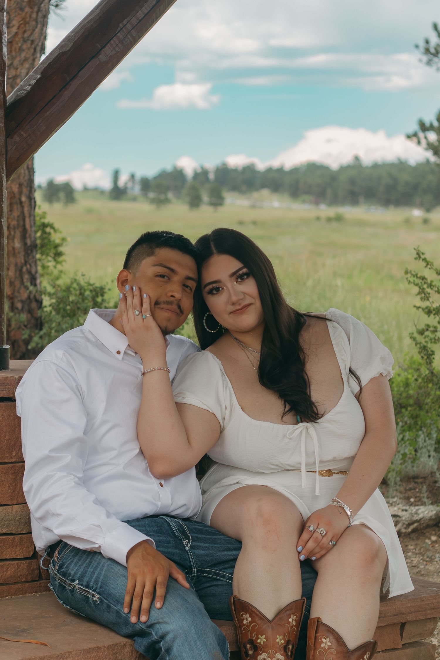 A couple in western-inspired outfits sits together on a wooden porch with mountains in the background.