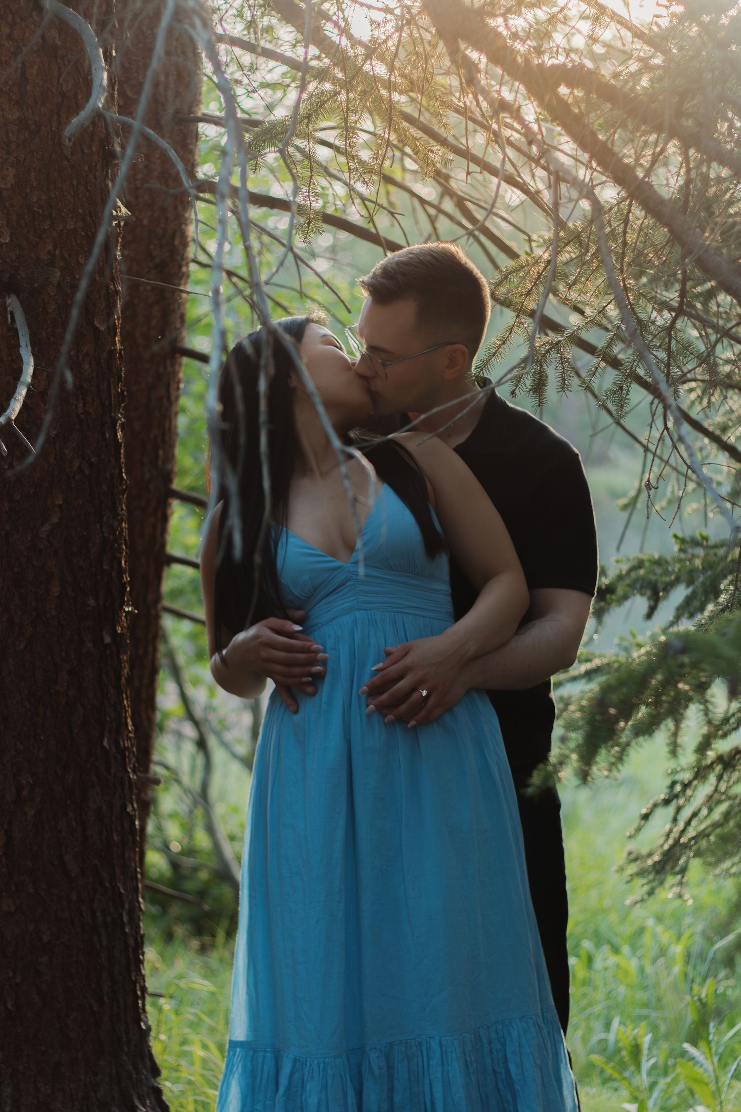 Couple in intimate pose against forest backdrop with sunlight filtering through branches.