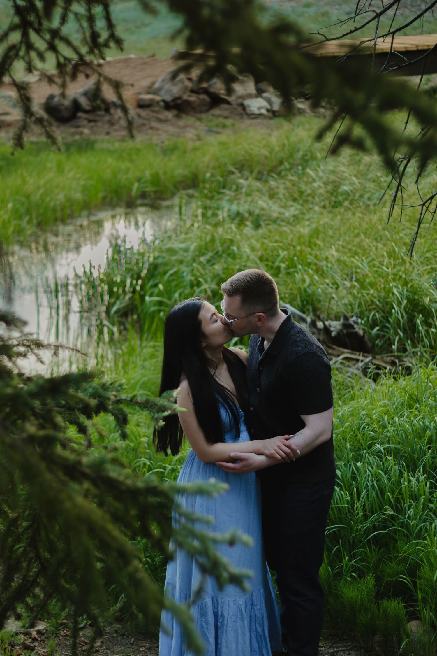 A couple embracing by a pond surrounded by lush green grass and trees.