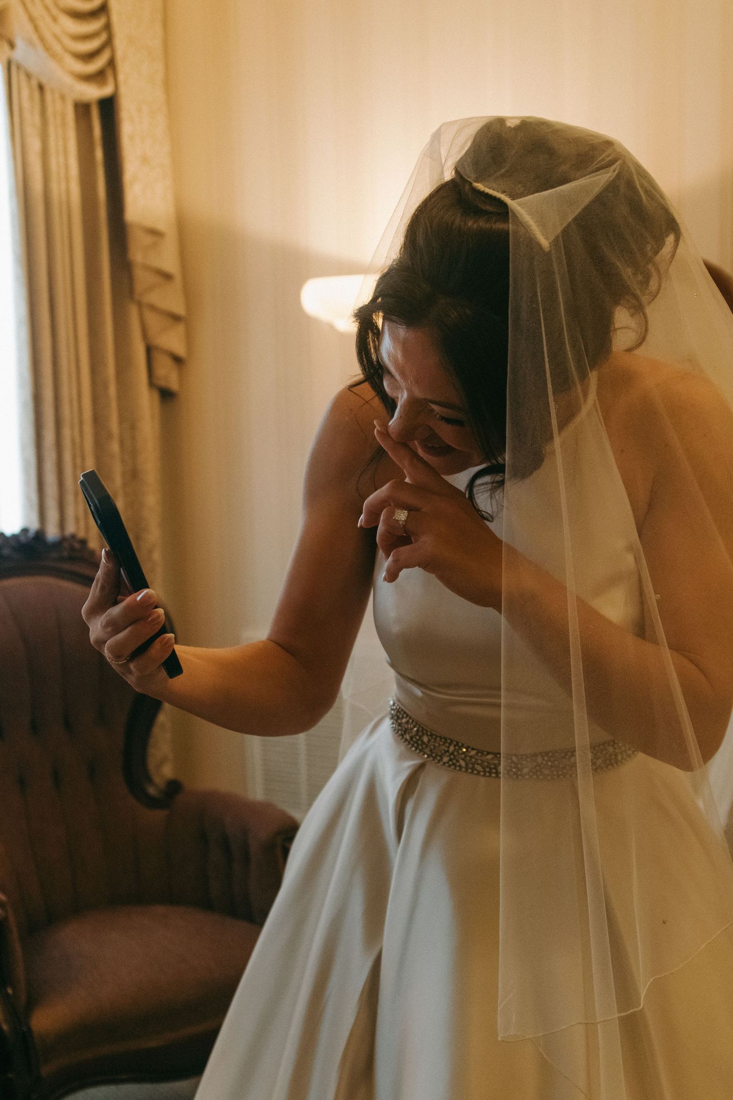 A bride in a white wedding dress with a beaded waistband and veil stands in a warmly lit room.