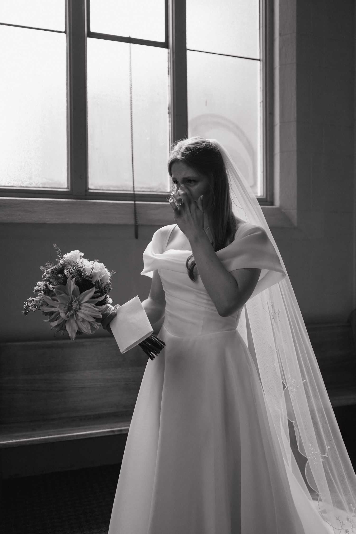 Emotional bride in white gown and veil holding bouquet, wiping tears in black and white wedding photo.