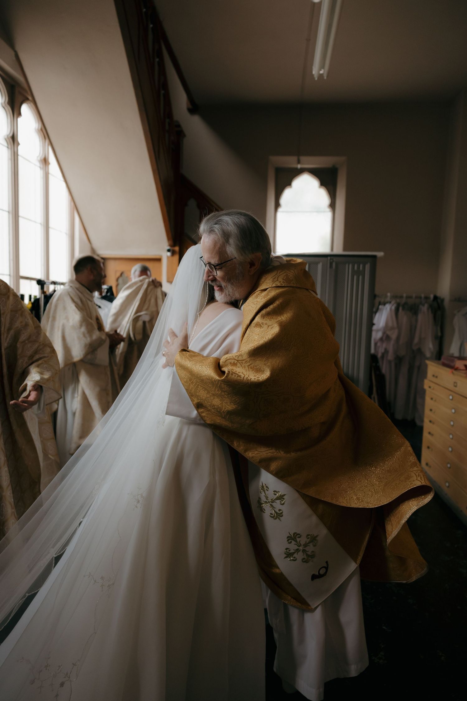 Priest in gold vestments embracing bride in white wedding gown and veil in church vestry before ceremony.