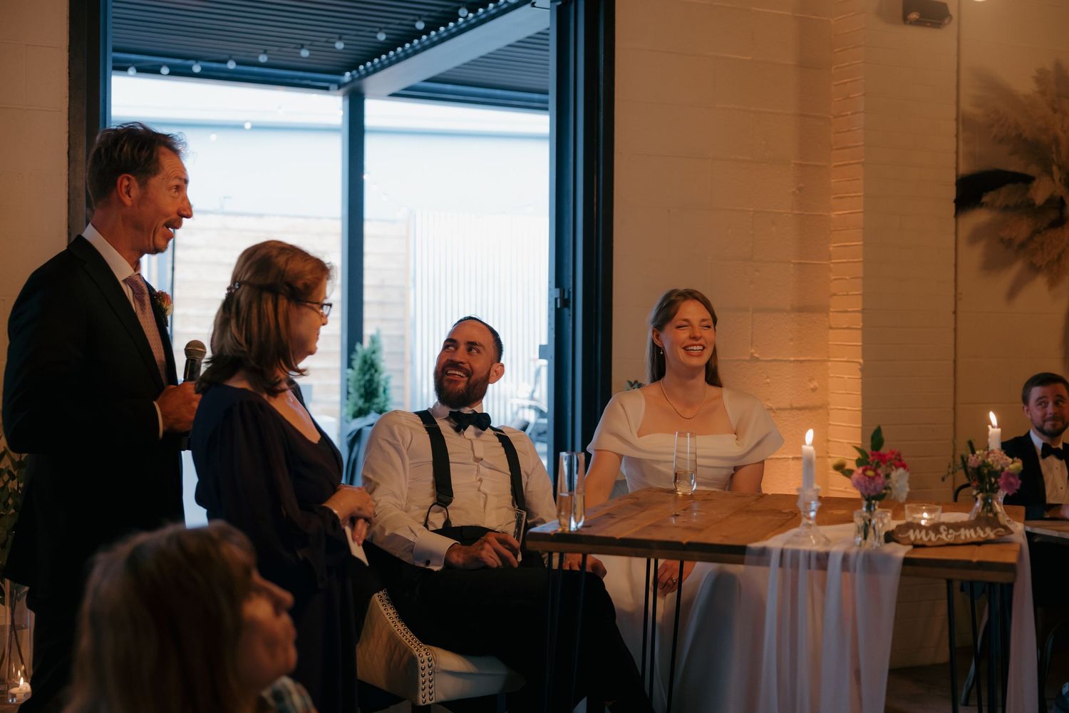Father giving a toast to smiling bride and groom seated at reception dinner table.