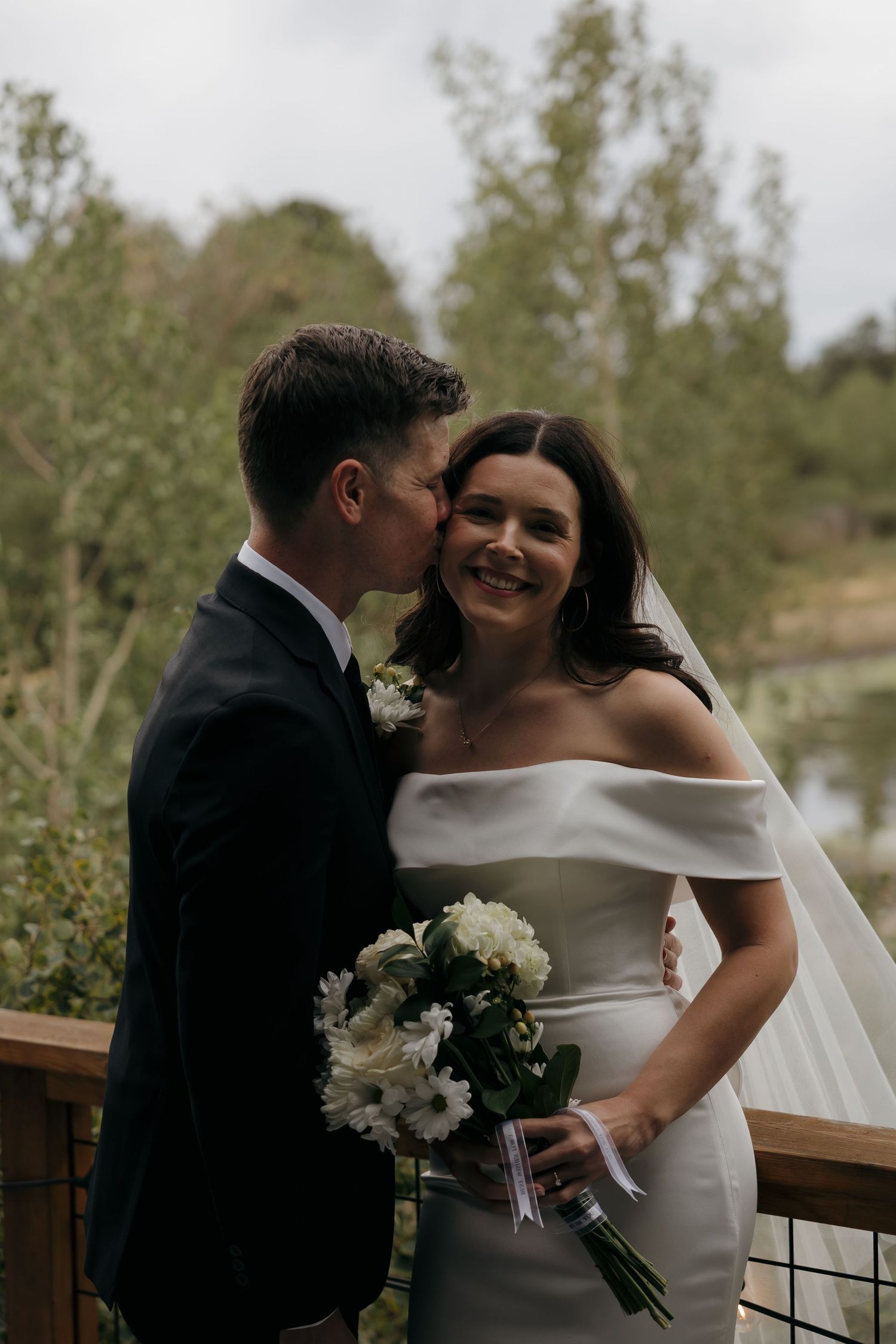 Groom kissing bride on cheek as she smiles holding white floral bouquet outdoors.