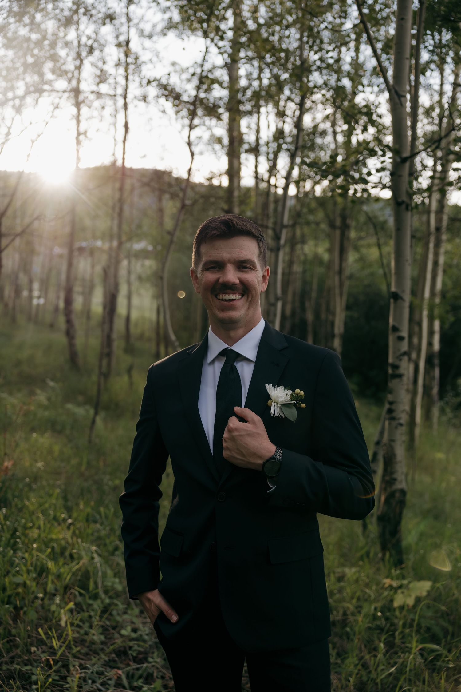 Smiling groom in a dark suit with boutonniere posing confidently in a sunlit forest during wedding portraits.