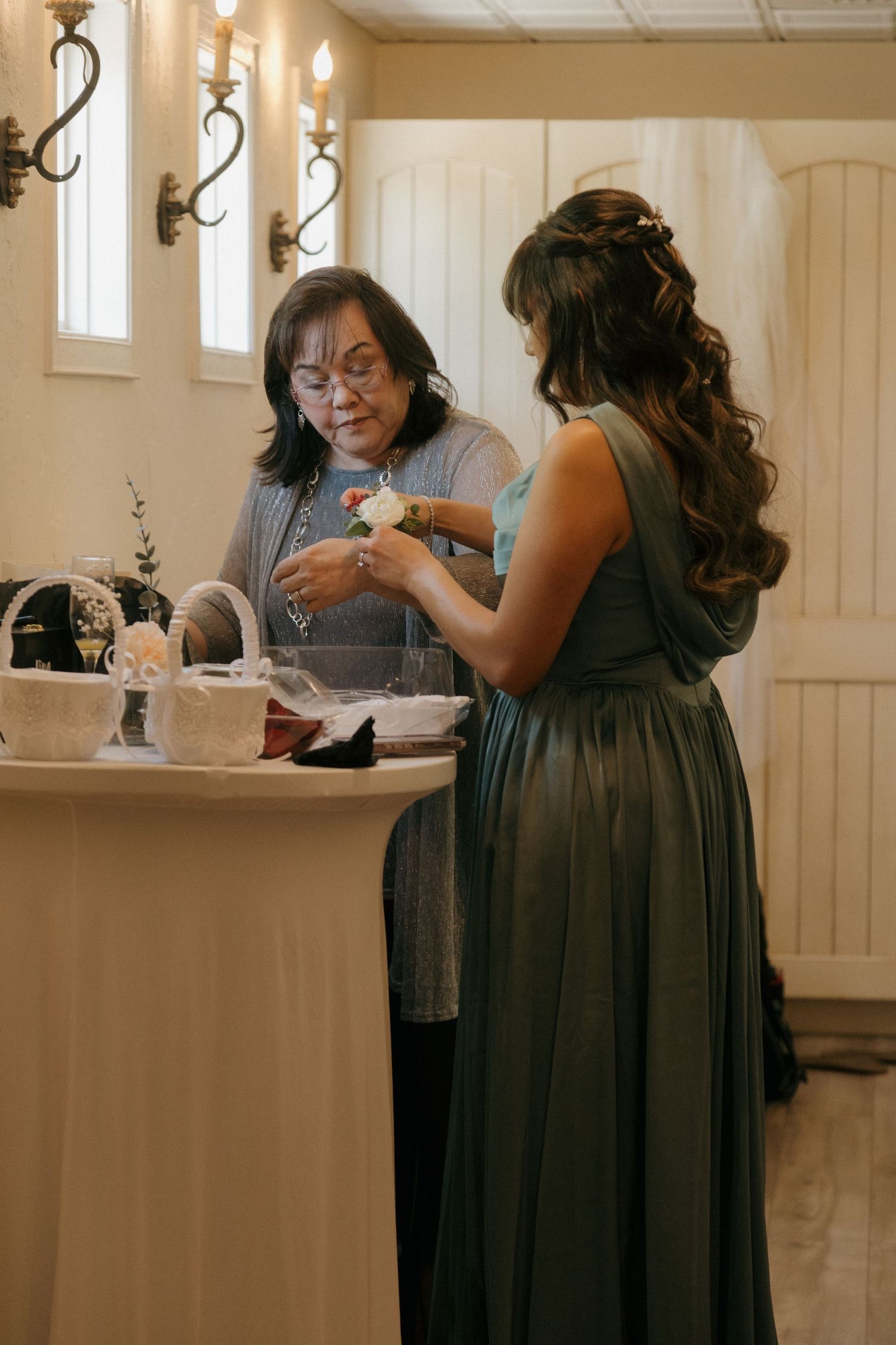 Woman helping a bridesmaid in a green gown pin a white boutonniere at a wedding venue.