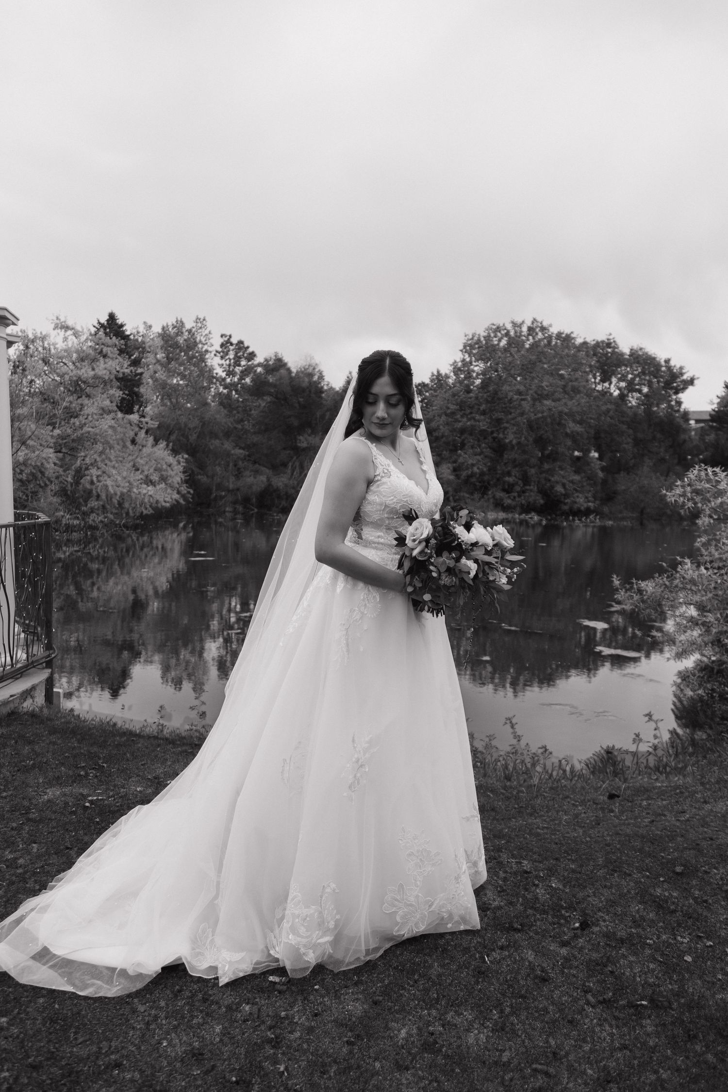 Black and white portrait of bride in white gown holding bouquet beside a reflective lake.