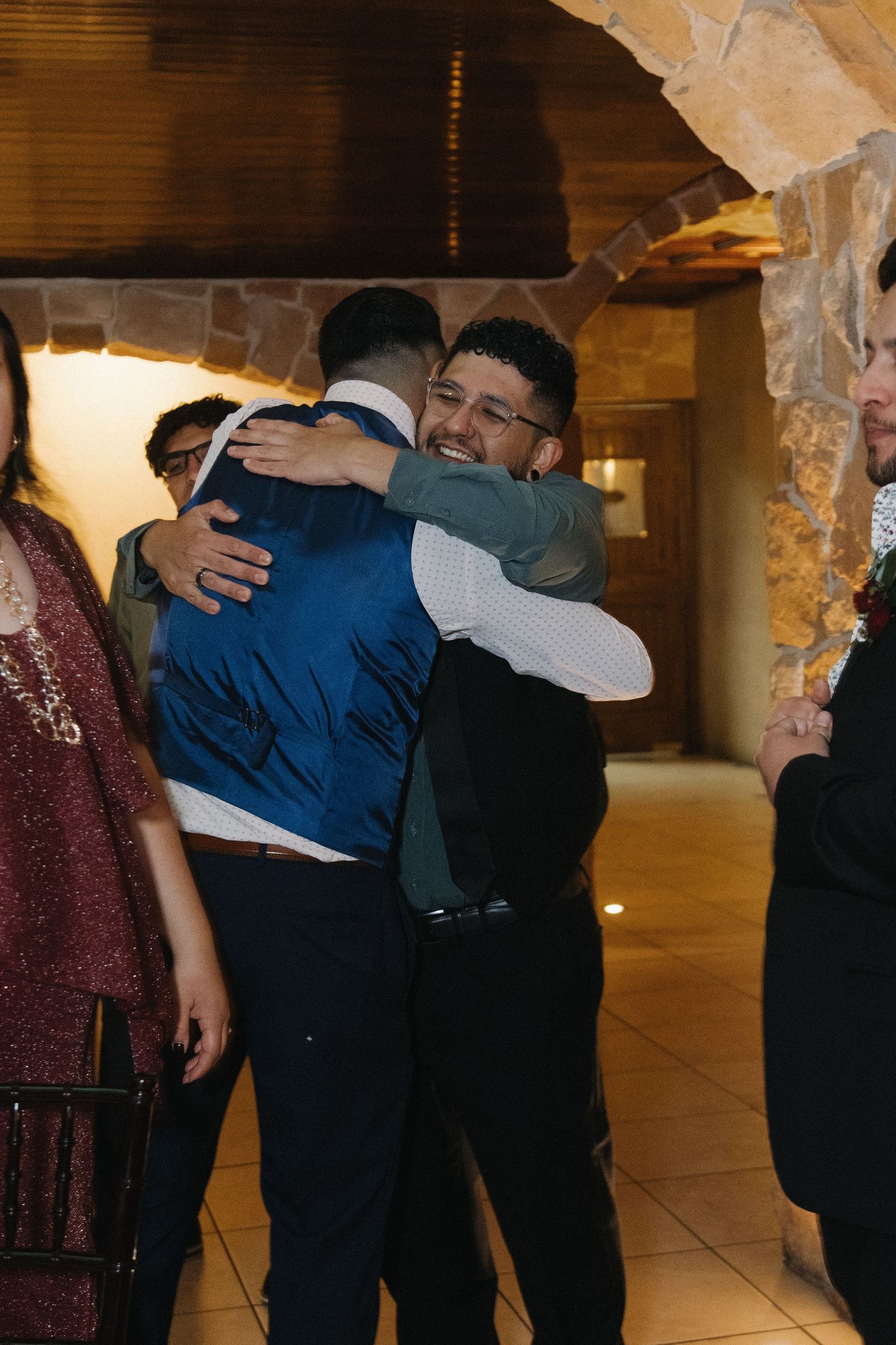 Two men share a warm hug at wedding reception in stone-walled venue with guests nearby.