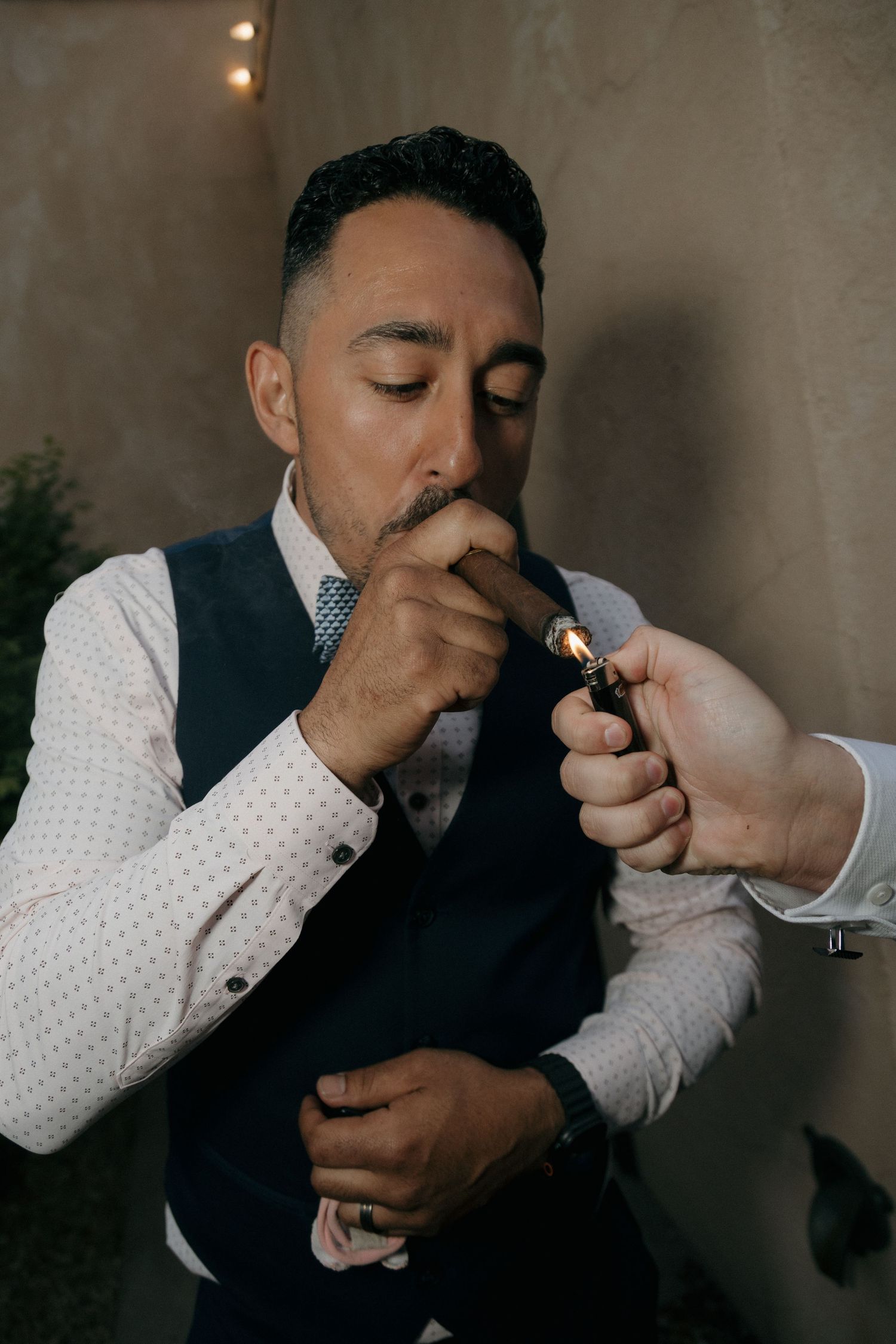 Elegantly dressed man in vest lights a cigar with a golden lighter at a wedding reception.