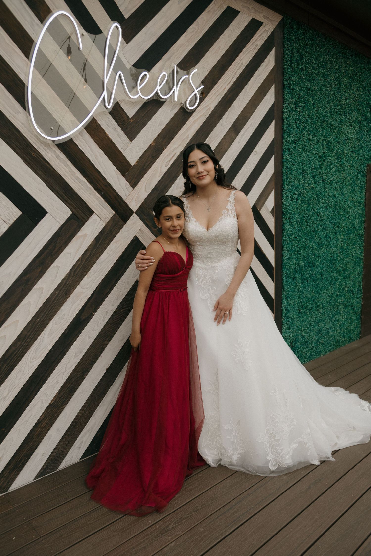 Bride in lace gown poses with young girl in red dress before geometric black and white wall.