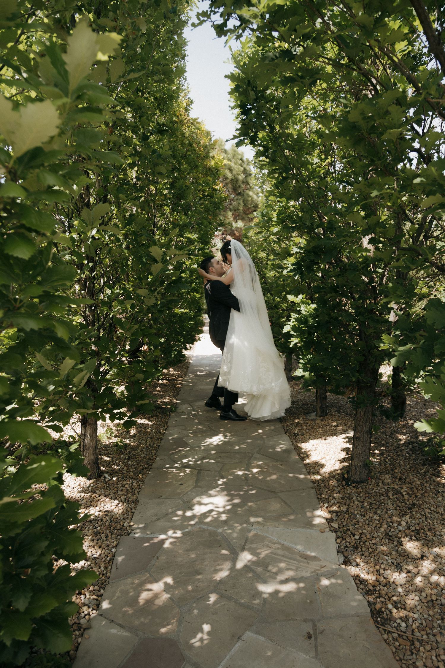 Bride and groom share a romantic kiss on a stone garden pathway lined with lush green trees on their wedding day.