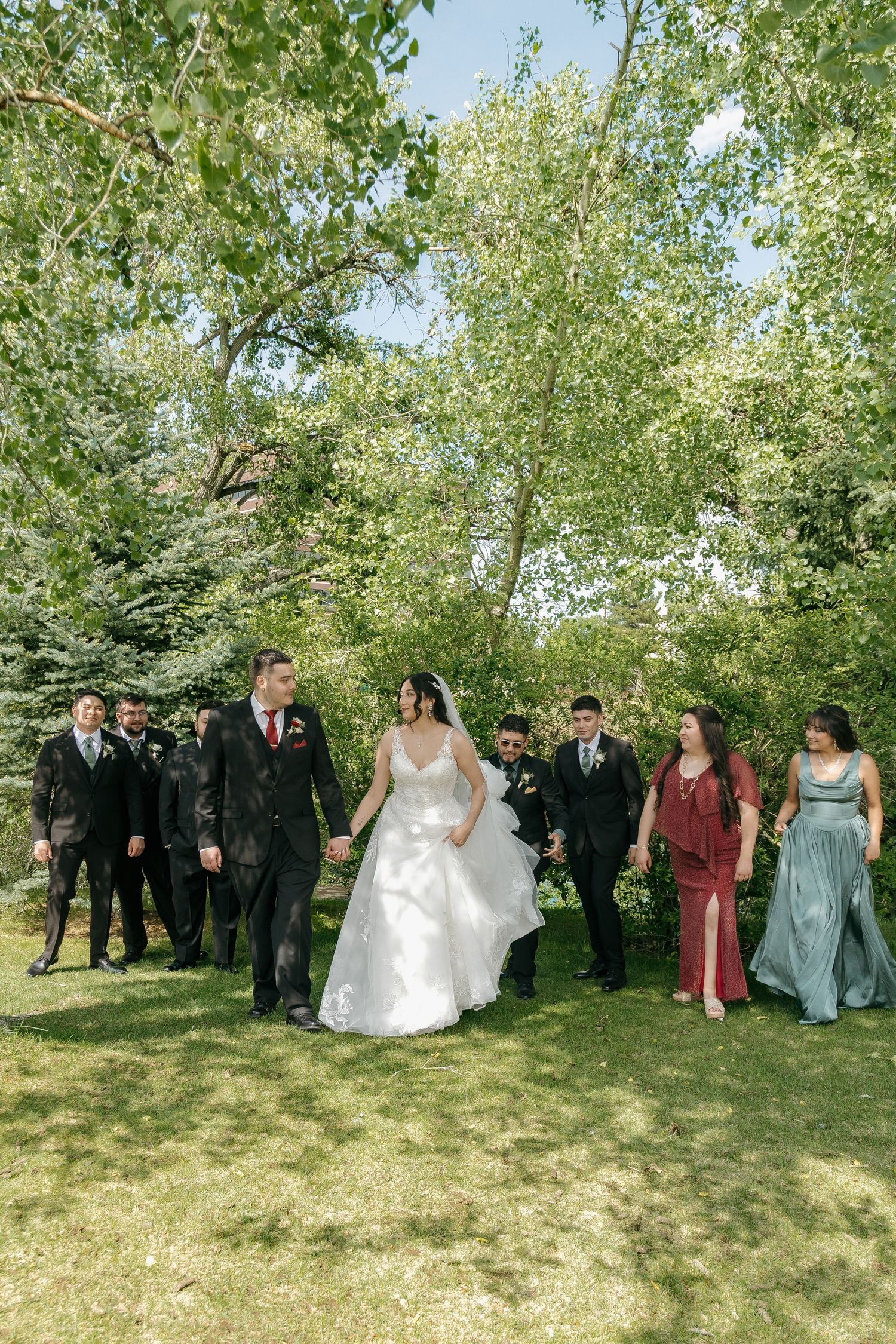 Wedding party walks through lush green garden, bride in white gown with groom and attendants in formal attire.