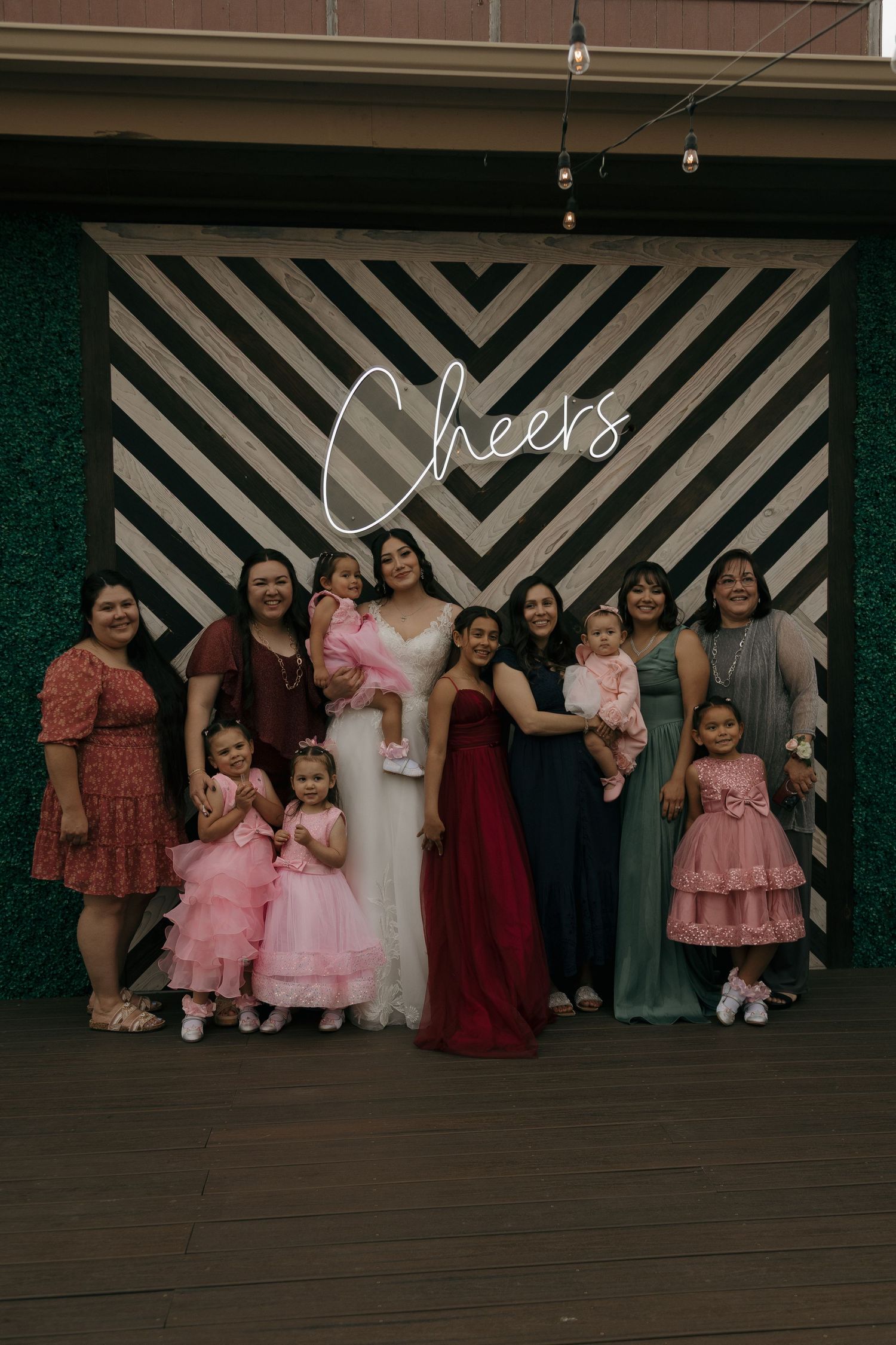 Bride poses with female guests and young children in front of a striped Cheers neon sign backdrop.