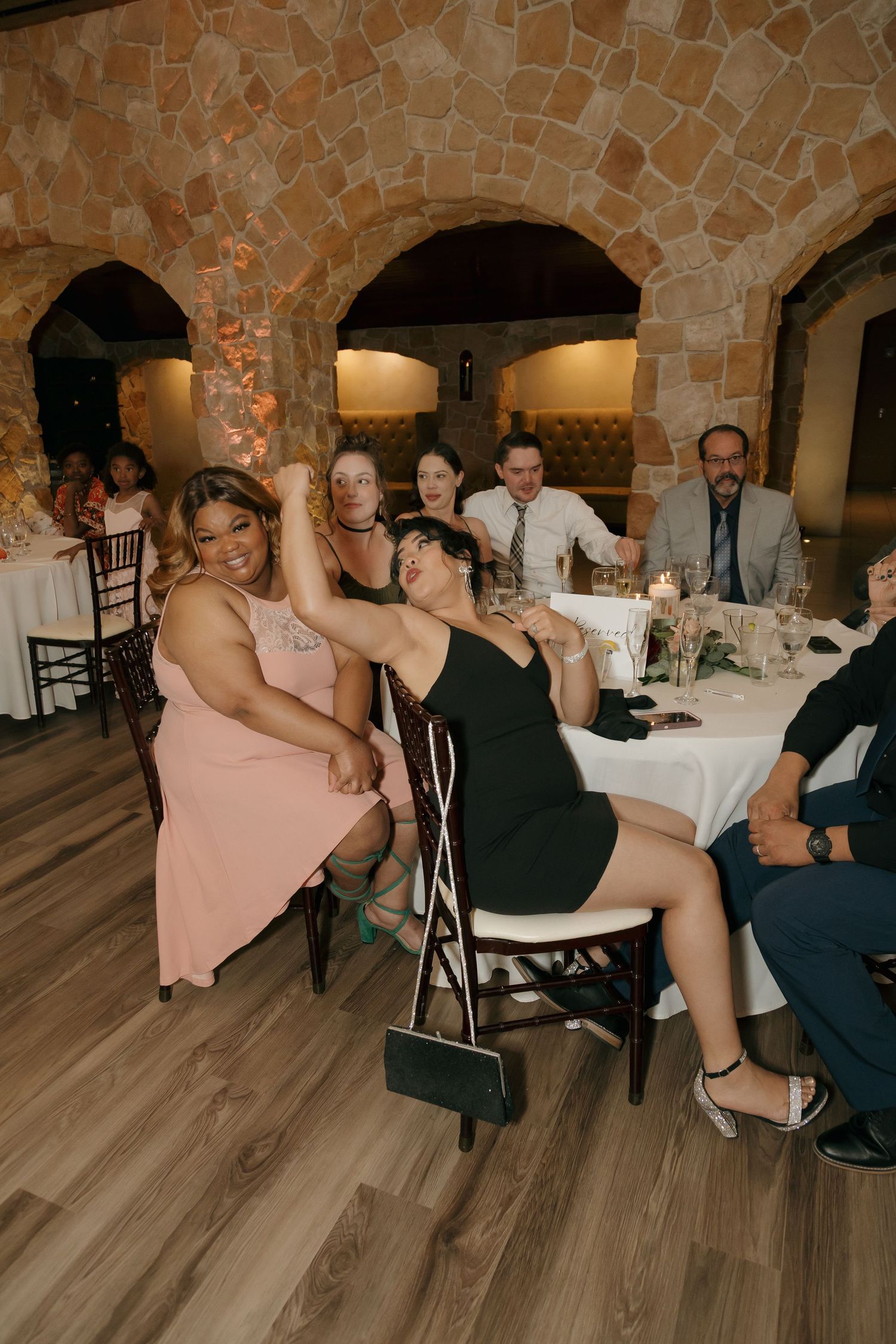 Wedding guests laughing and posing at reception table in rustic stone venue.