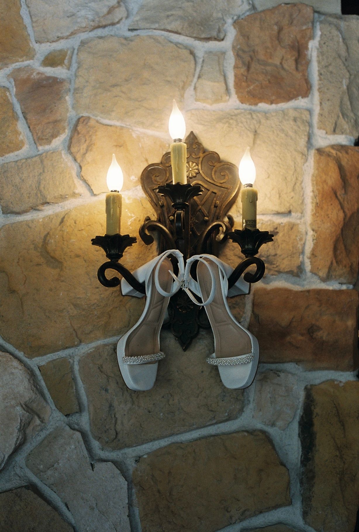 White bridal heels hanging on ornate iron candelabra wall sconce with lit candles against rustic stone wall.