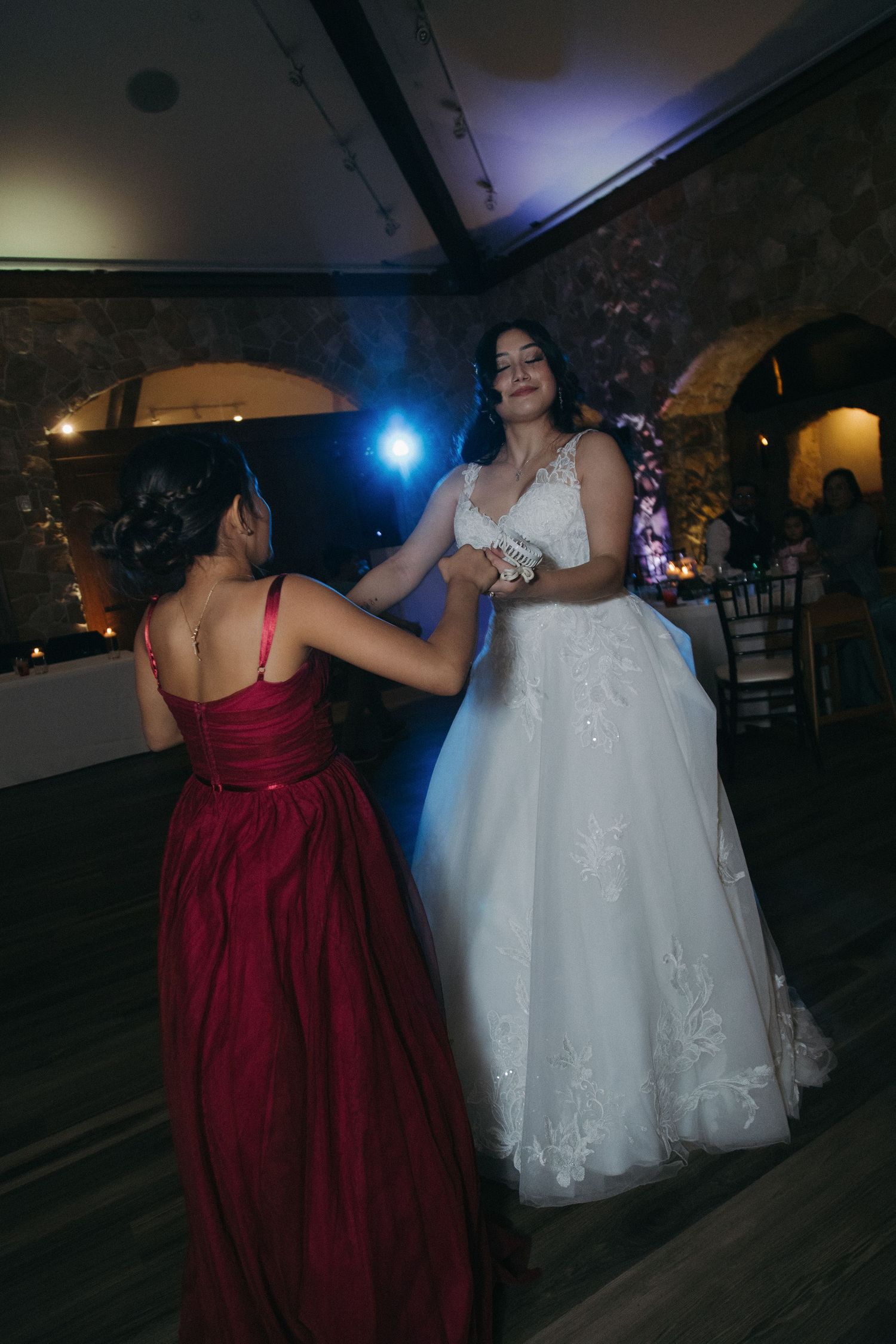 Bride in white lace gown dancing with guest in red dress at wedding reception.