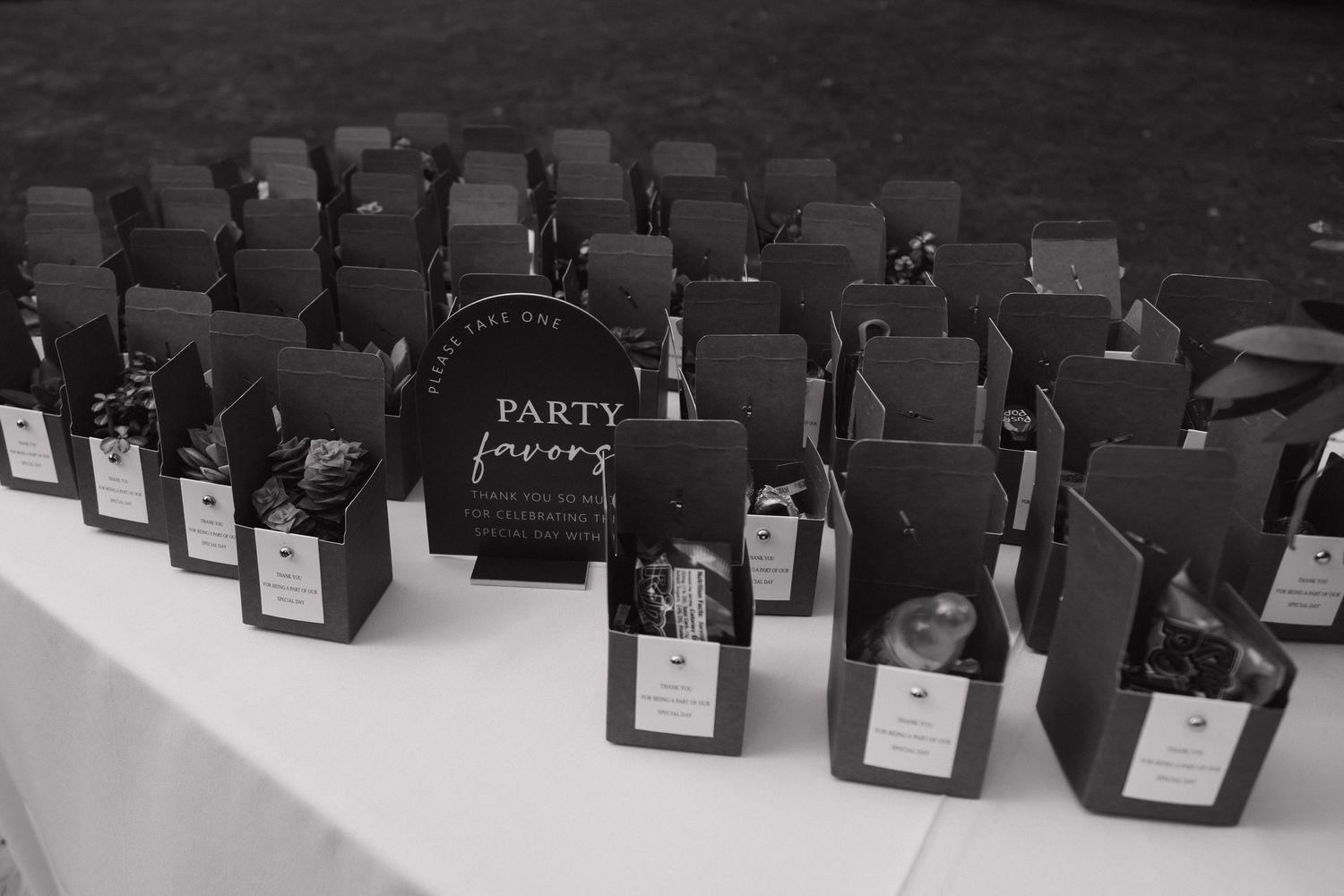 Black and white photo of elegant party favors displayed on a white table at an outdoor event.