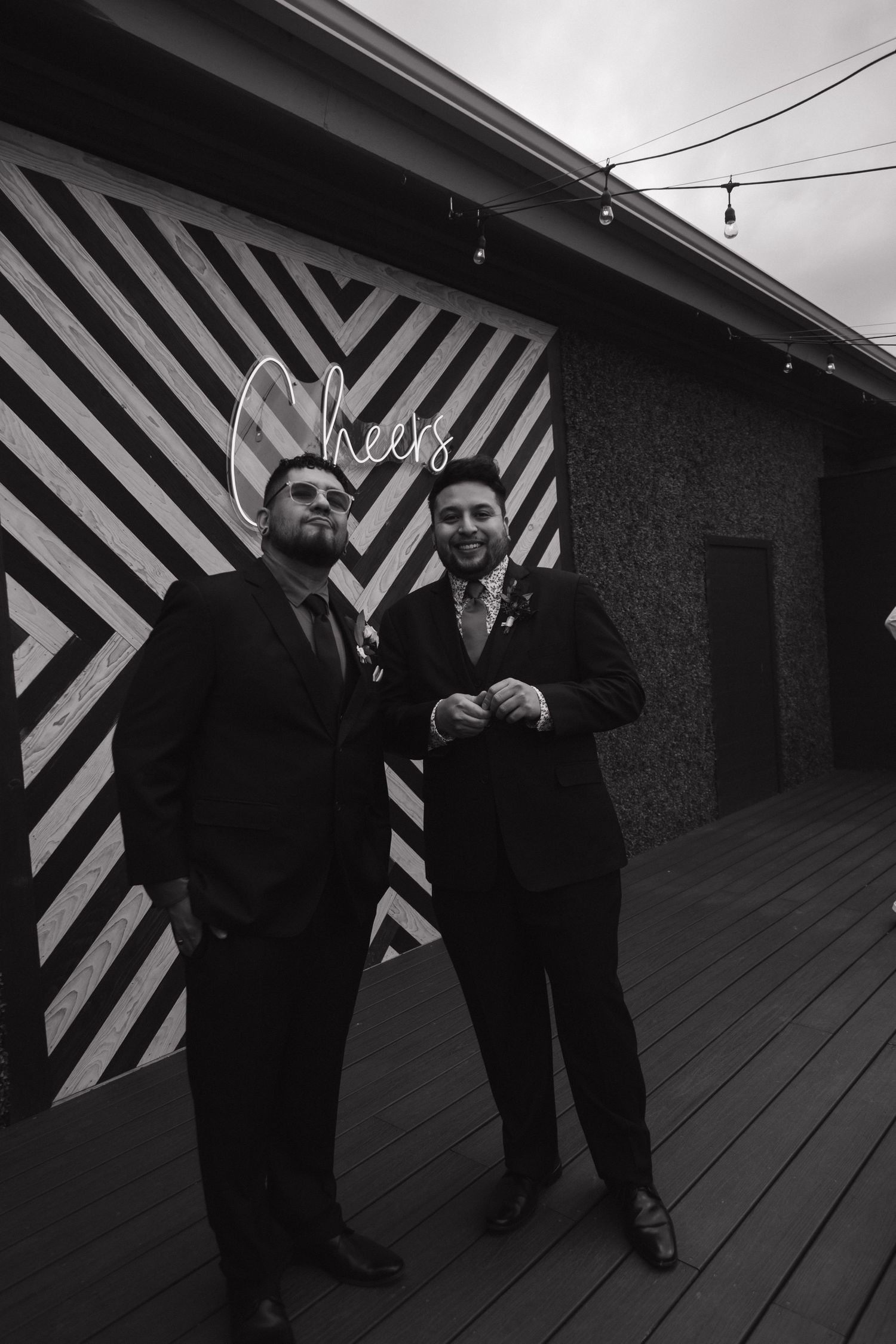 Two groomsmen in black suits pose together outside venue with decorative Cheers sign in black and white photo.