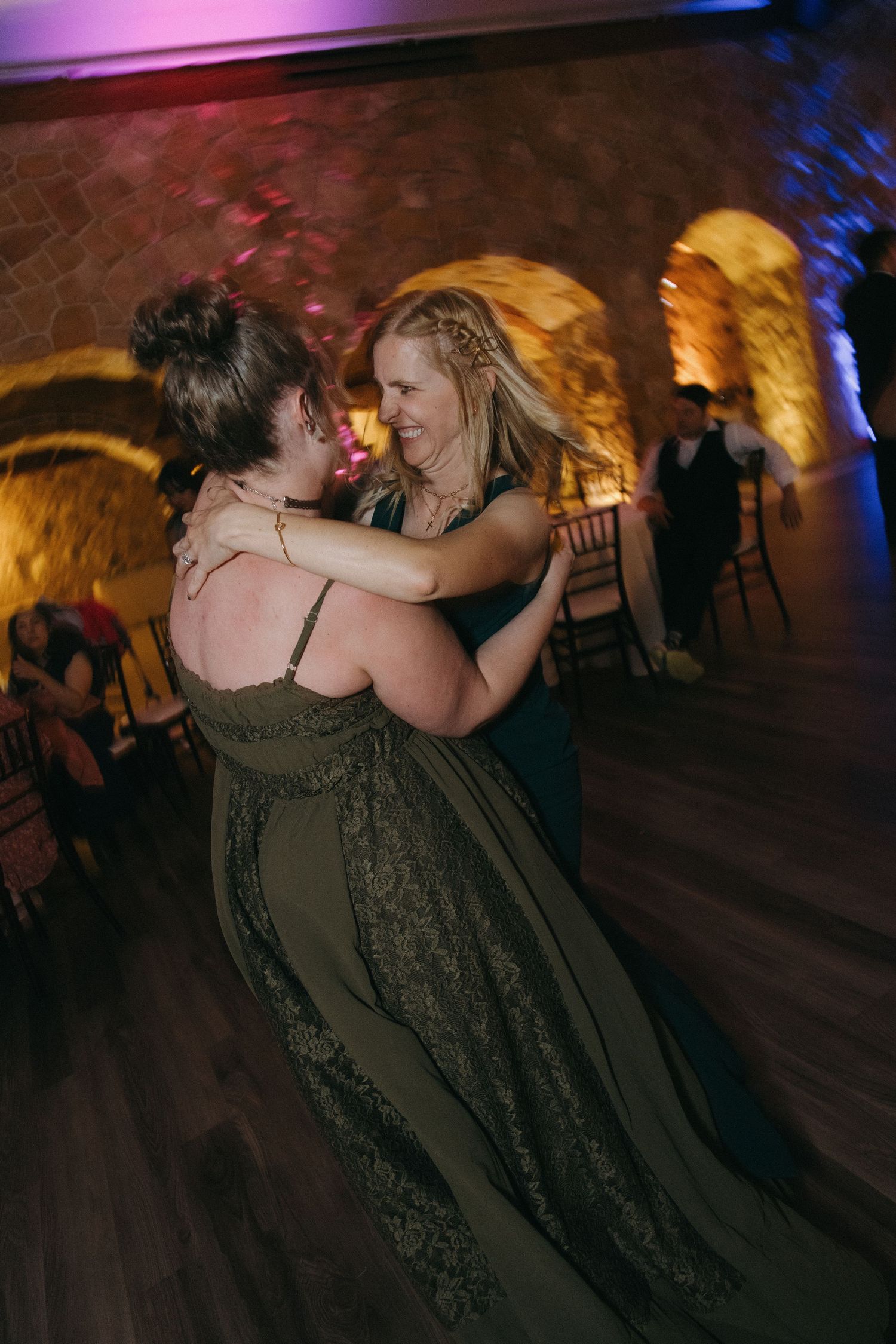 Two women in elegant olive green gowns share a warm embrace while dancing together at a wedding reception.