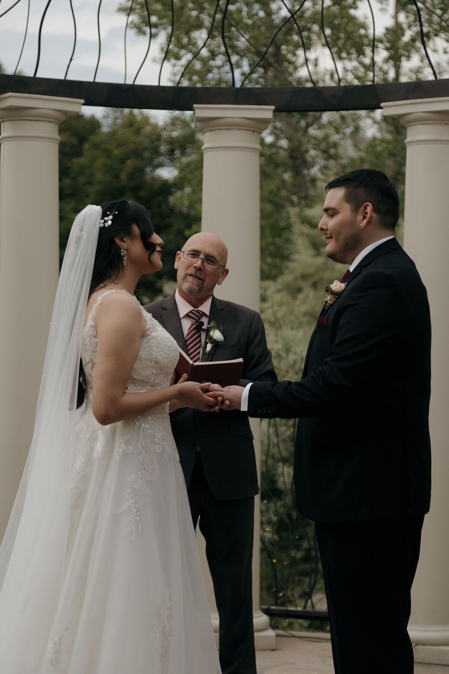 Bride and groom exchange vows holding hands before an officiant under an elegant white column gazebo.