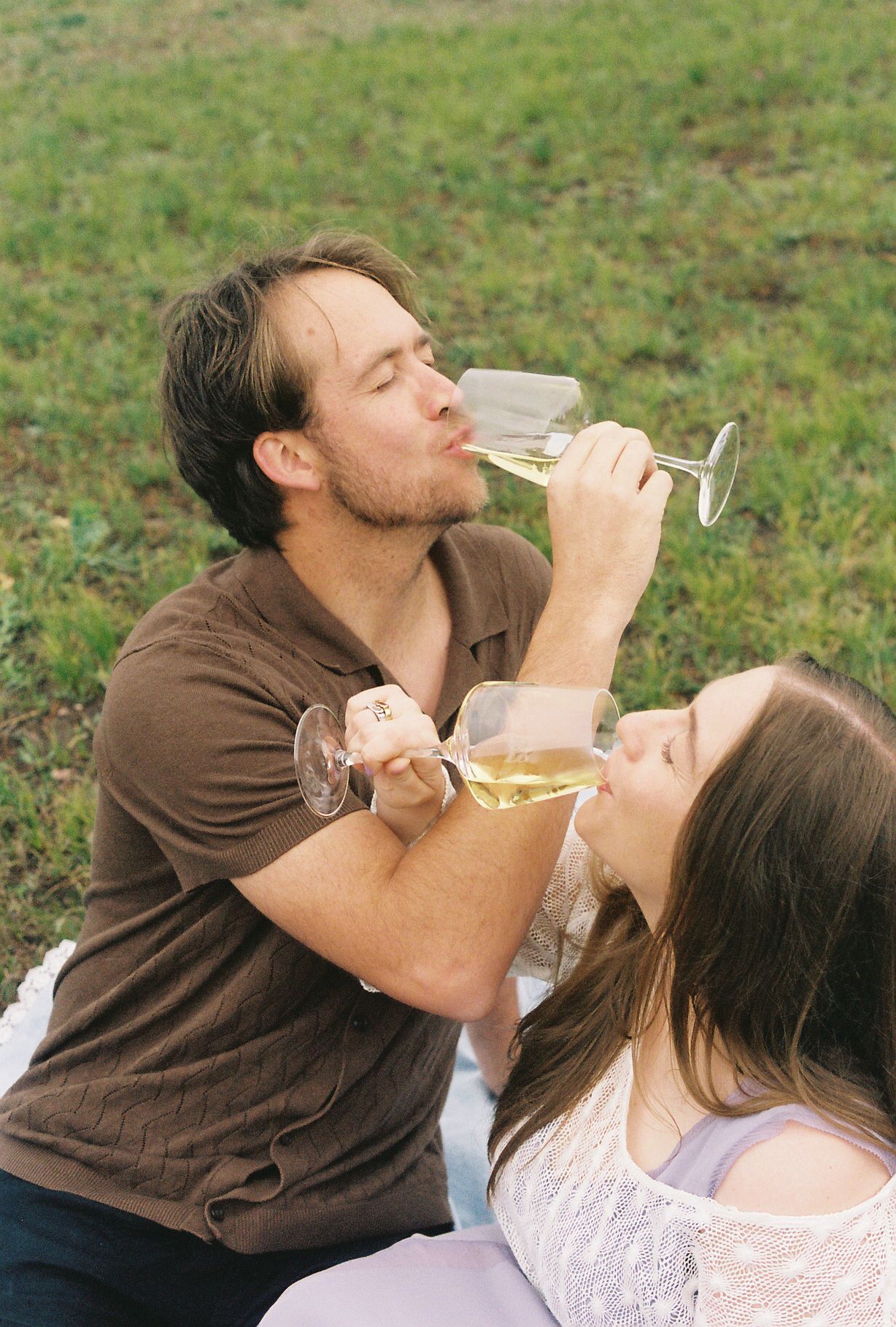 A couple shares wine glasses during a romantic outdoor picnic on a grassy lawn.