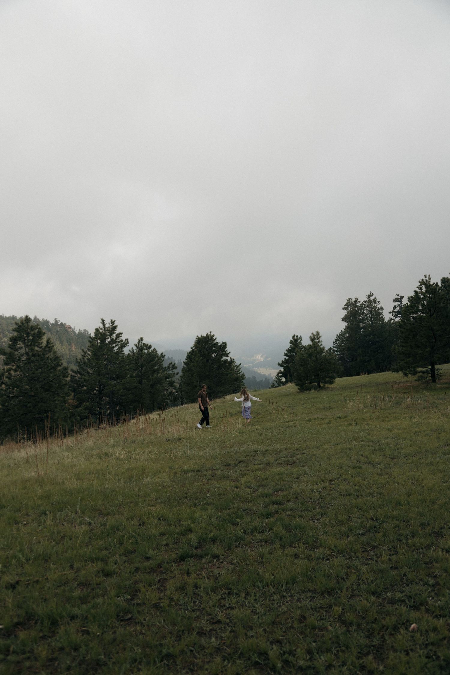 Couple running through misty mountain meadow surrounded by pine trees.