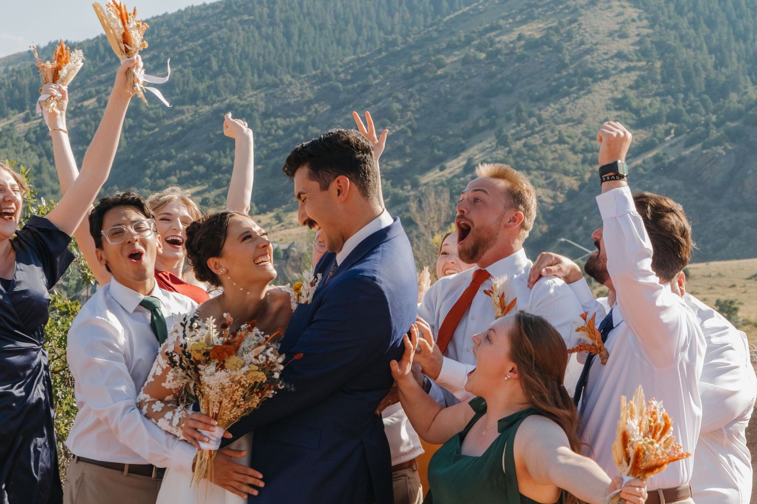 Wedding guests celebrate joyfully outdoors with raised hands against mountain scenery.