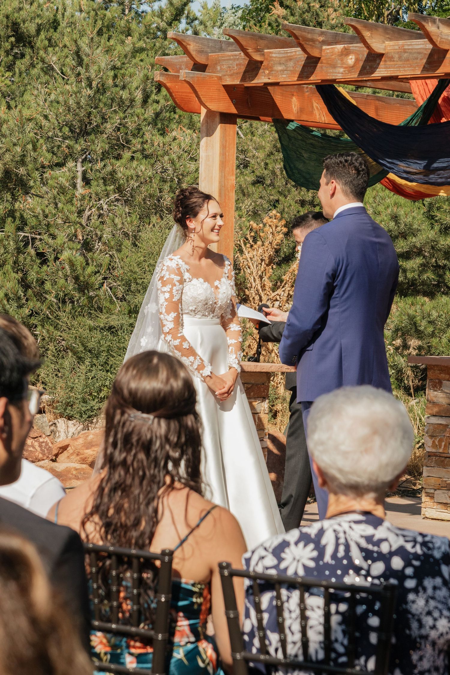Wedding guests watch ceremony under wooden pergola with natural desert landscape in background.