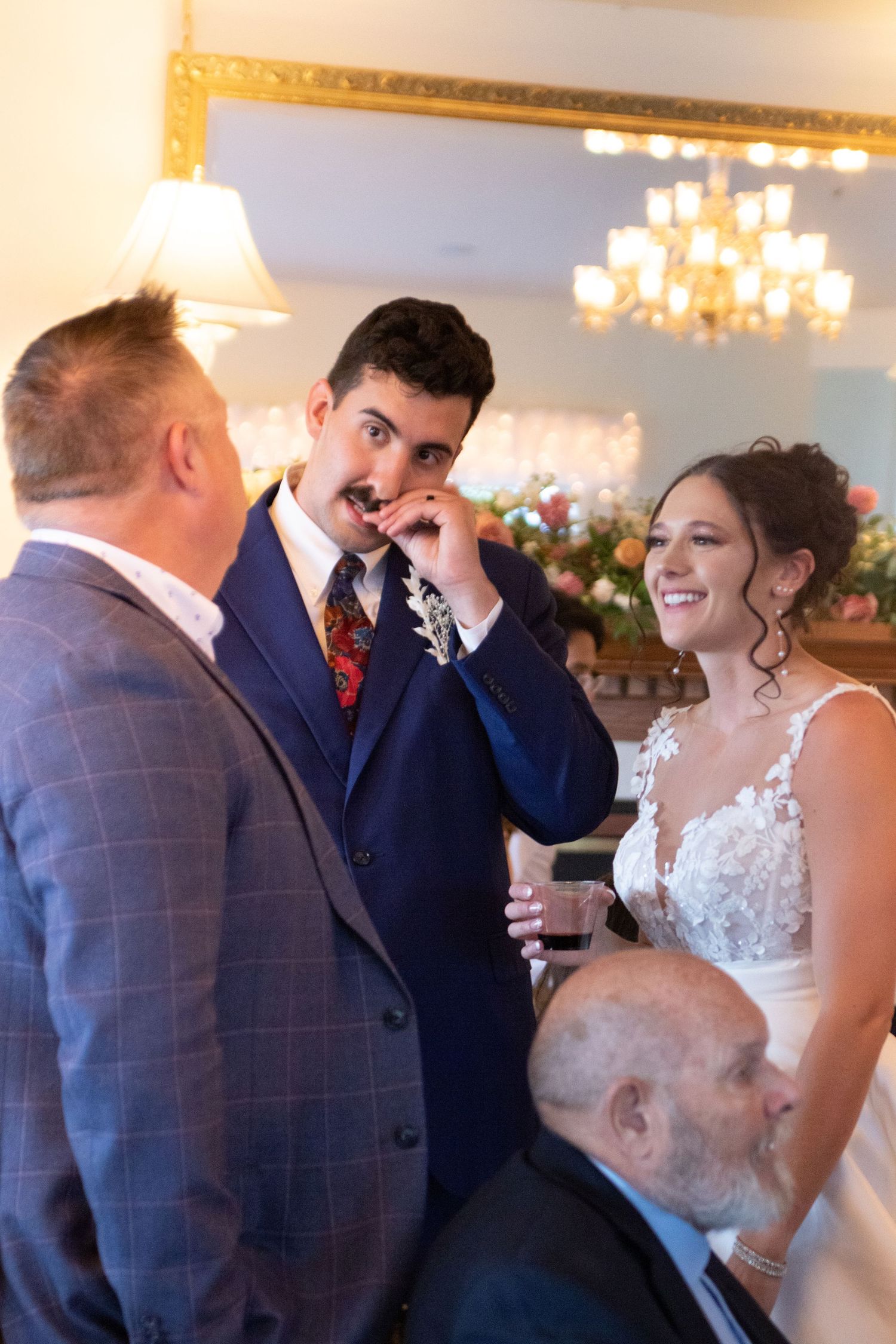 Wedding guests gather indoors under chandeliers during reception celebrations.