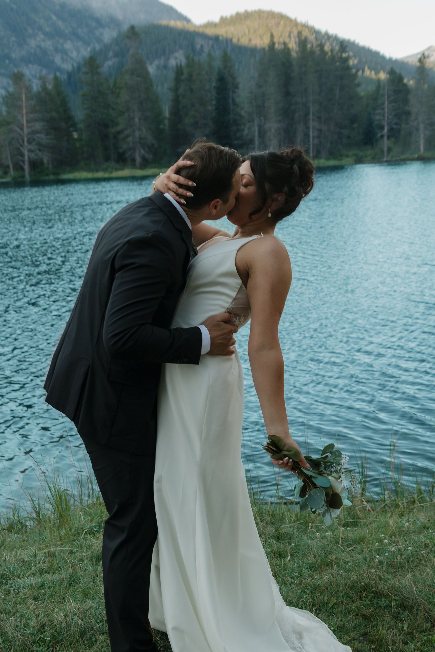 A romantic lakeside wedding moment unfolds with mountains and evergreens creating a stunning backdrop.