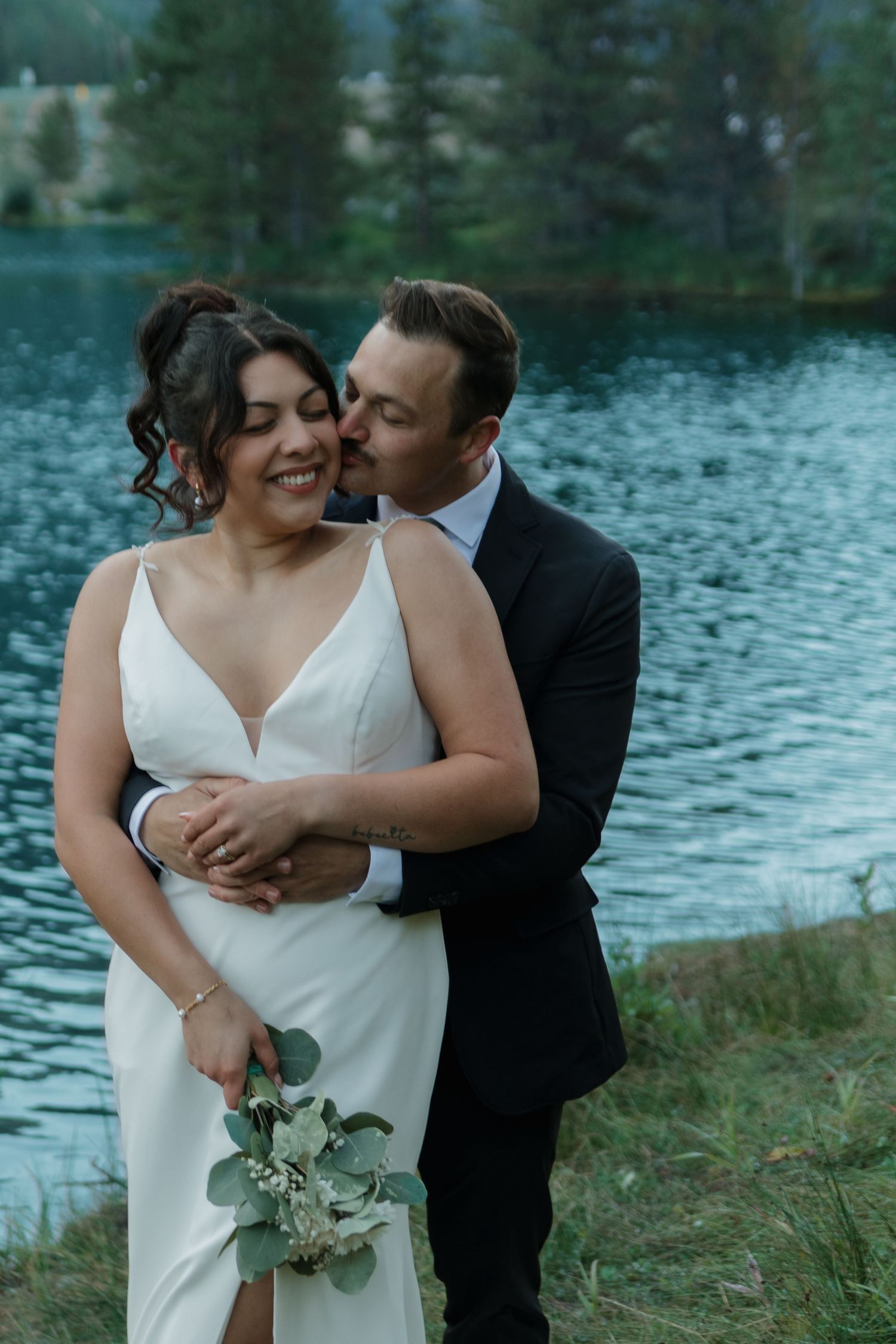A romantic lakeside wedding photoshoot with evergreen trees in the background.