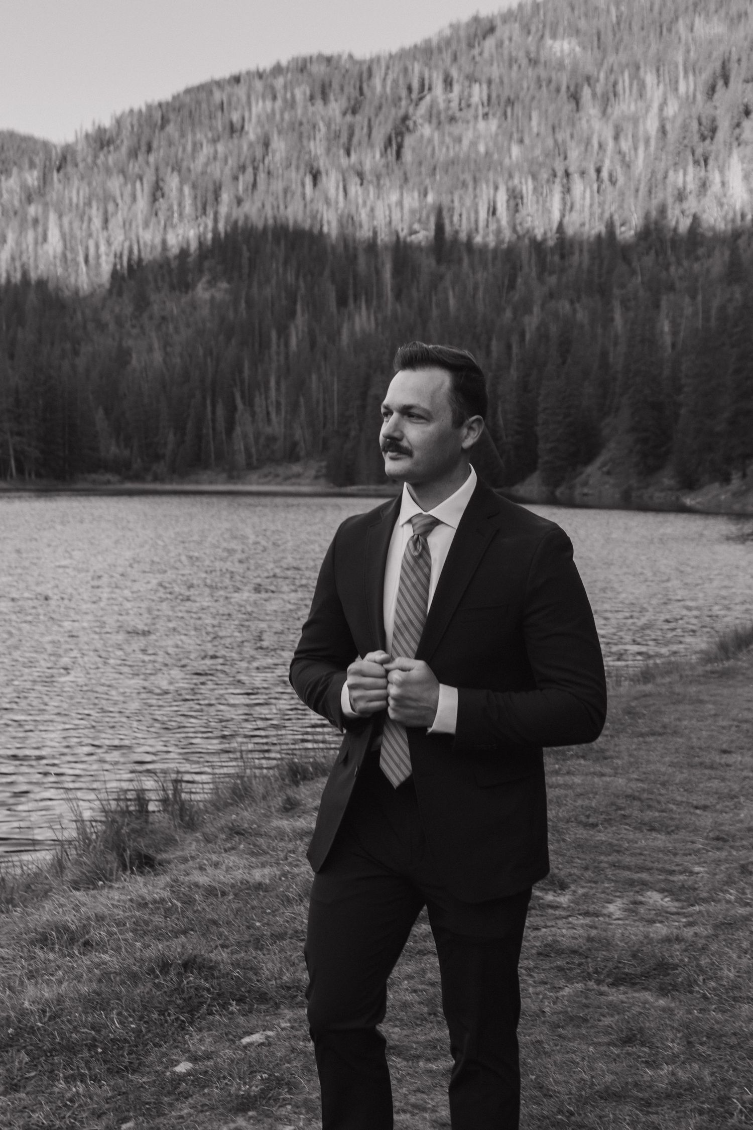 A figure in a dark suit stands against a scenic mountain lake backdrop in a moody black and white photograph.
