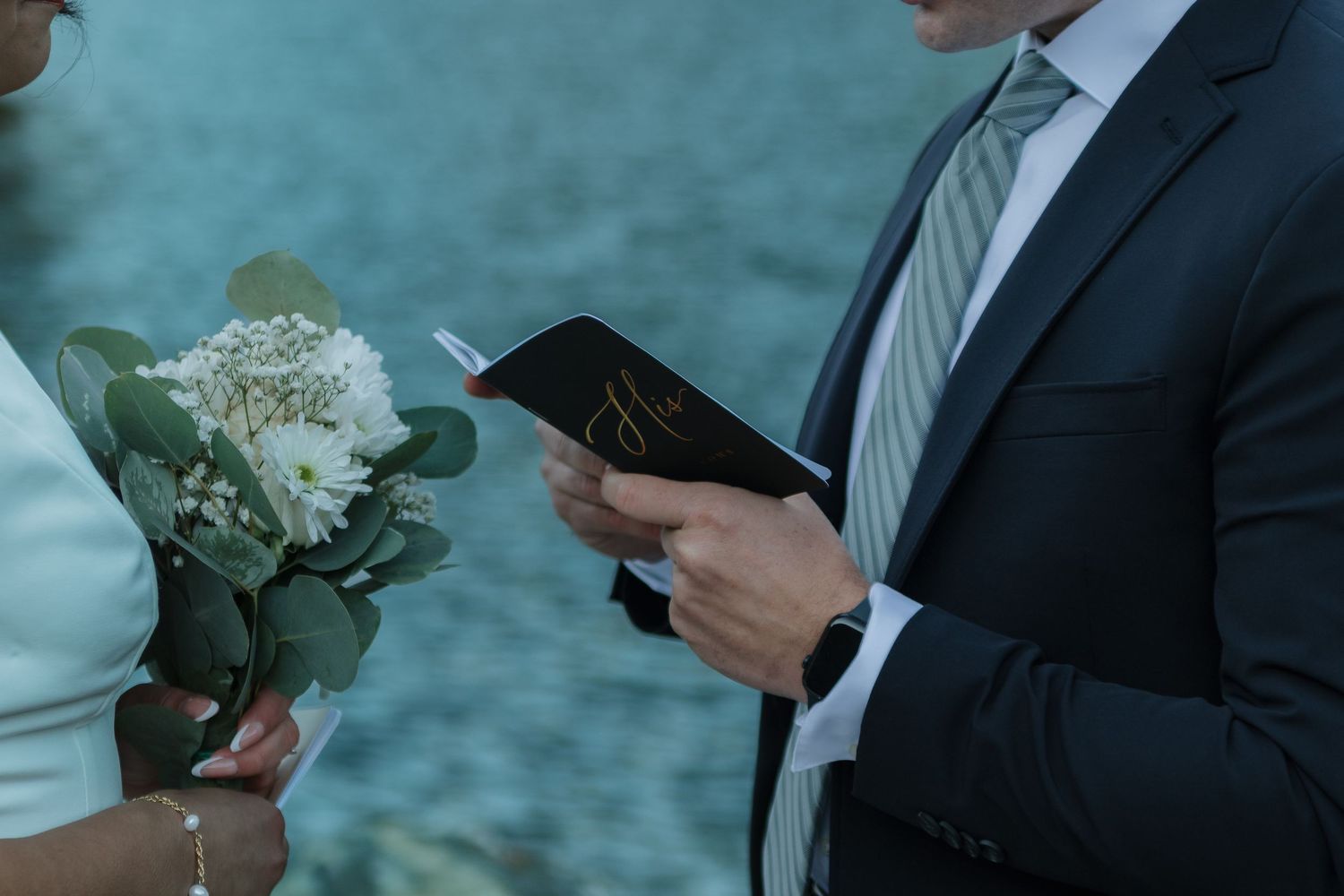 Two people in formal wear stand close together reading from cards beside a lake.