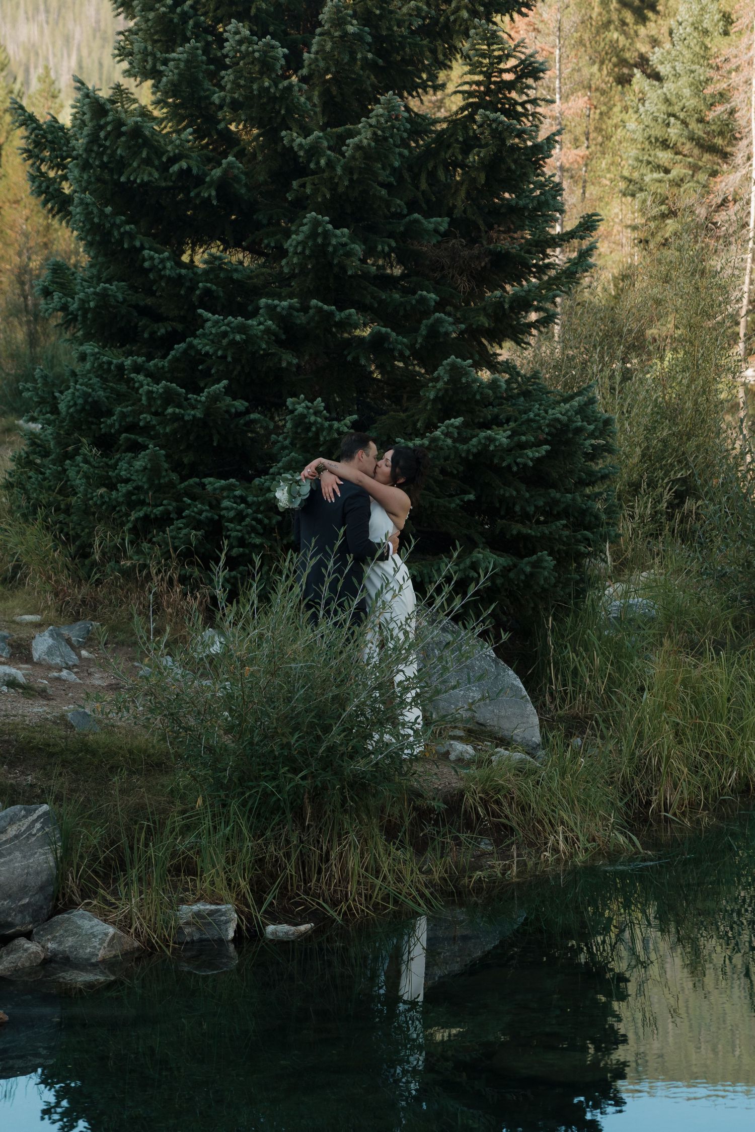A wedding couple poses near a serene mountain lake surrounded by evergreen trees.