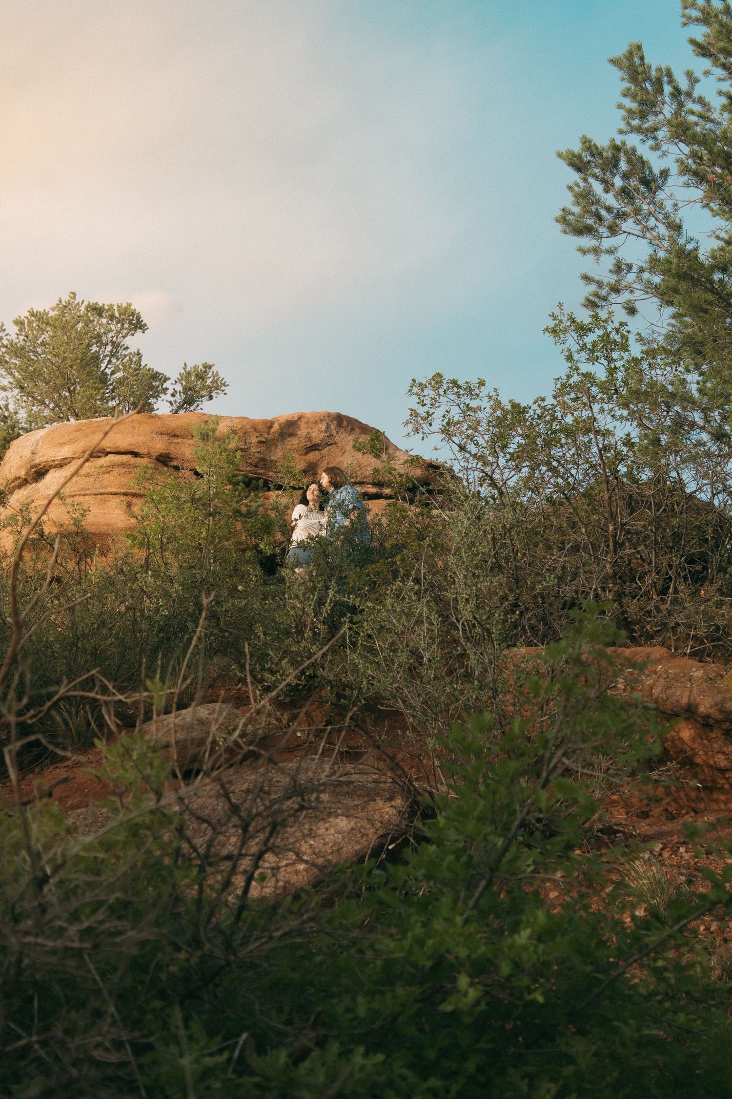 A distant view of two people standing on rocky terrain with trees and mountains in the background.