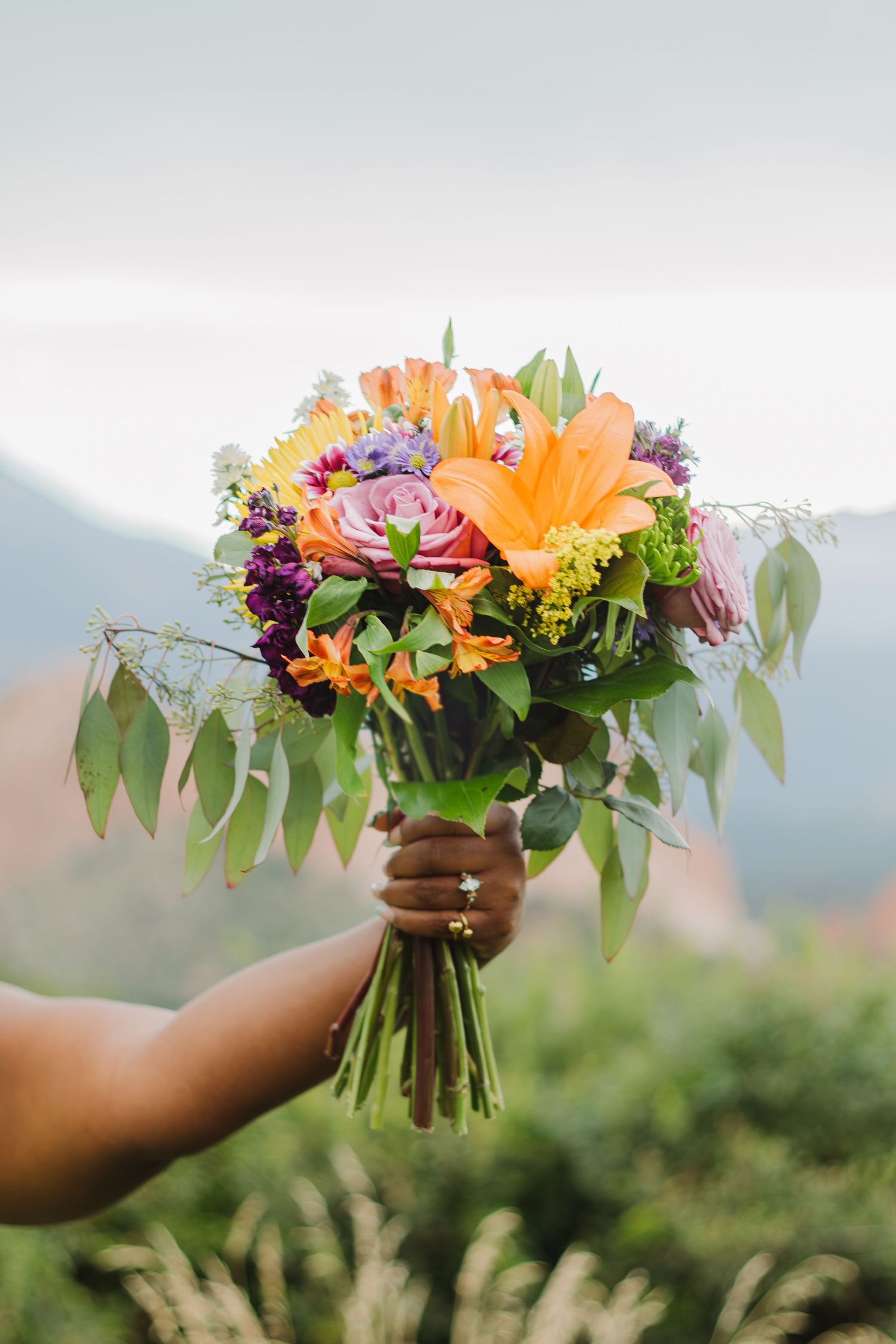 A vibrant bouquet of orange lilies, pink roses and purple flowers with eucalyptus leaves held against a mountain backdrop.