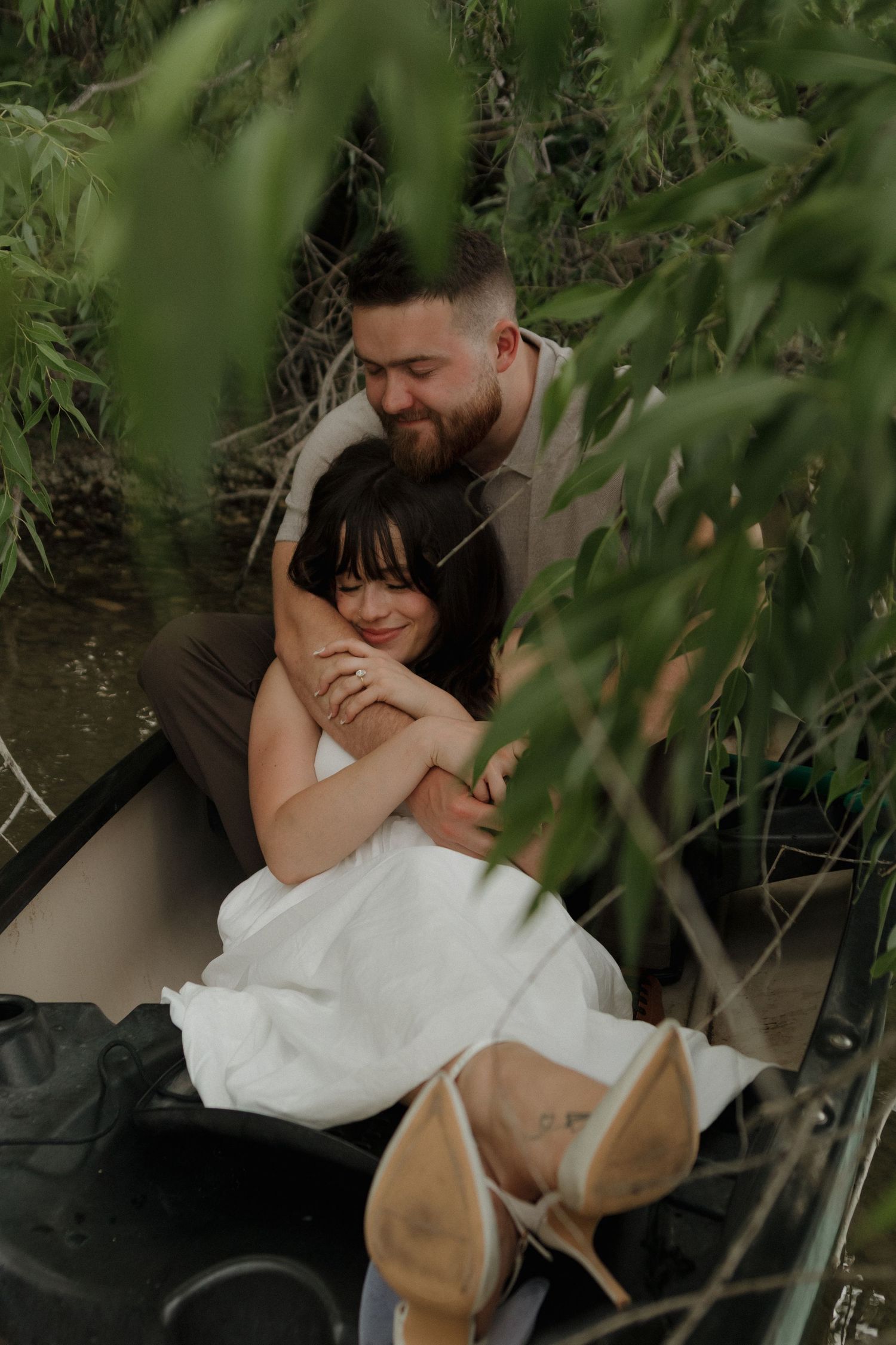 Two people share an affectionate embrace among green foliage by the water.
