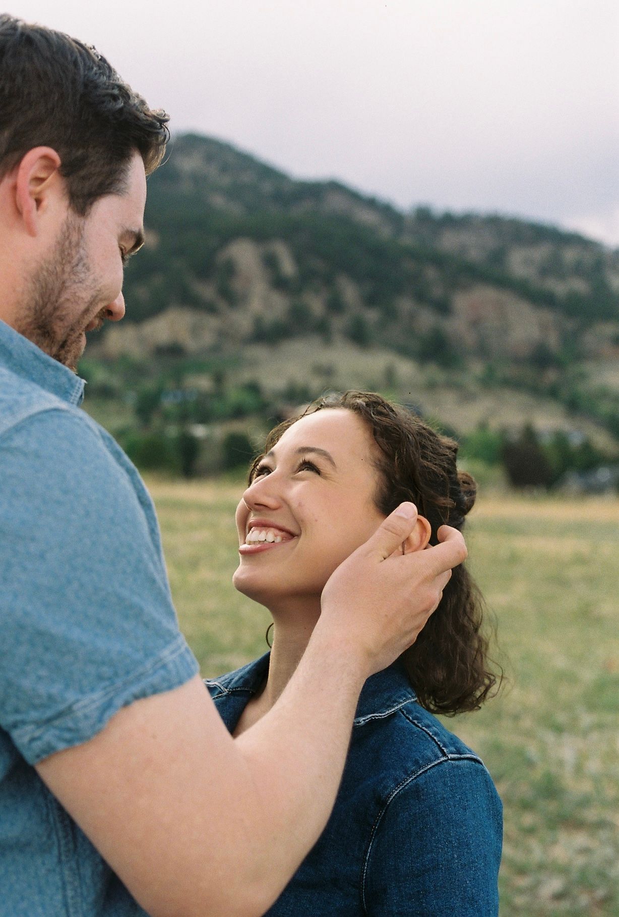 A couple shares a tender moment outdoors, man gently tucking woman's hair as she smiles up at him with mountains behind.