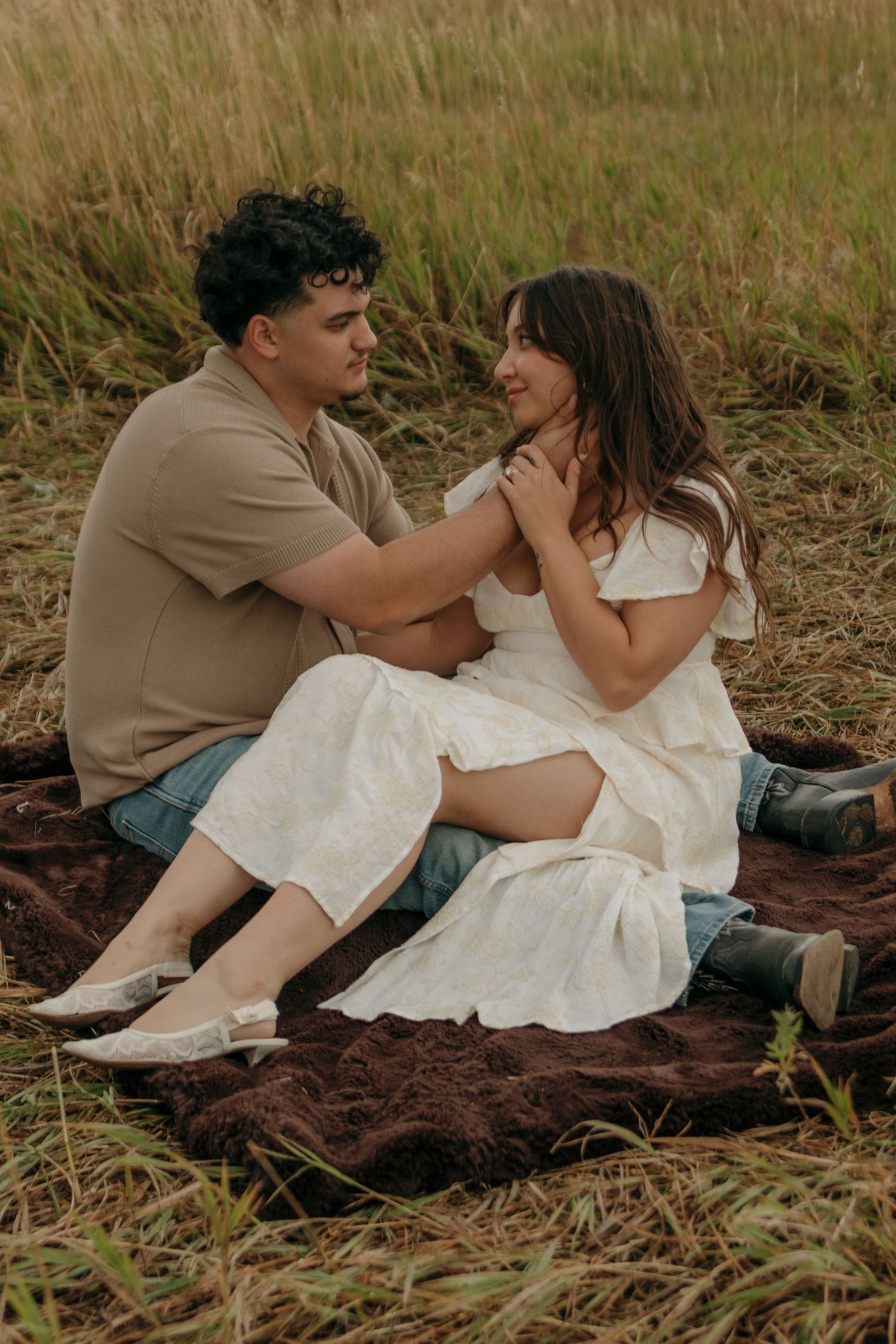 Couple sitting on brown blanket in grassy field, man holding womans hands while gazing at each other.
