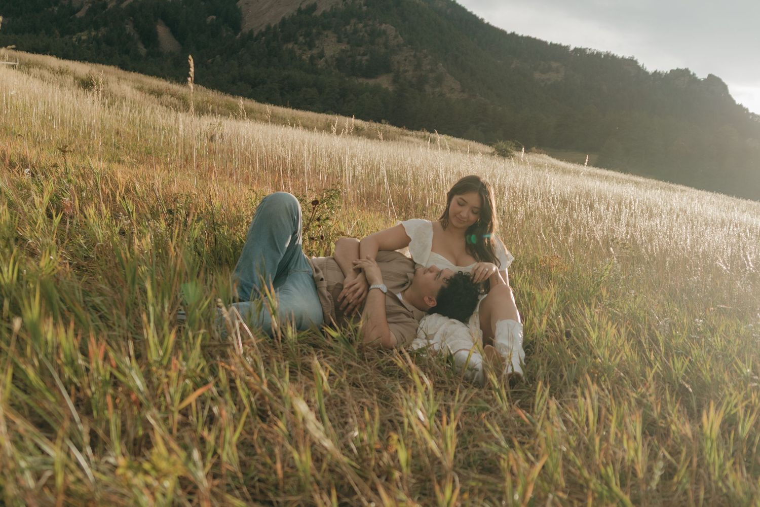 Couple relaxing together in a sunlit grassy meadow with mountains in the distance.