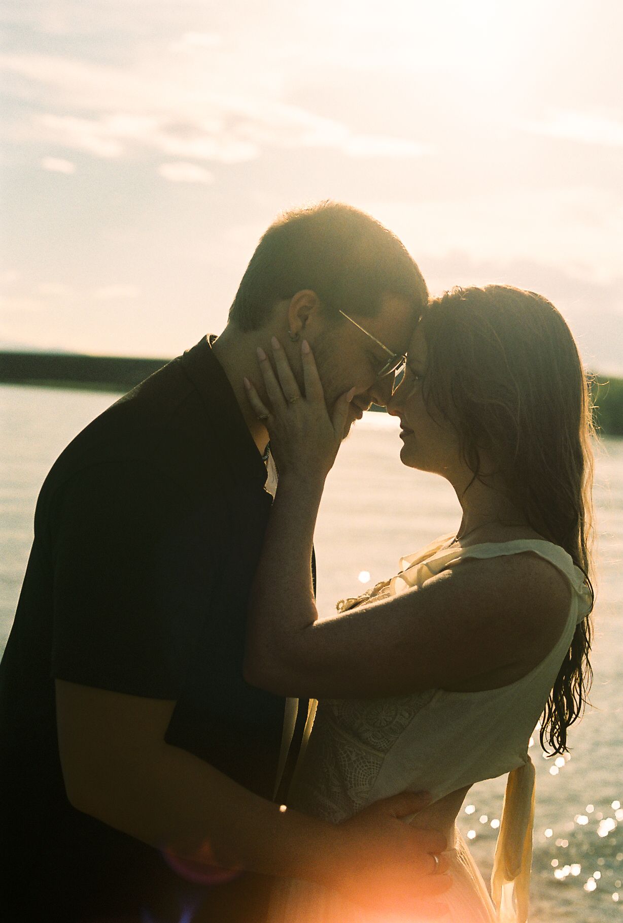Couple sharing intimate almost-kiss moment backlit by golden sunset glow near lakeside shore.