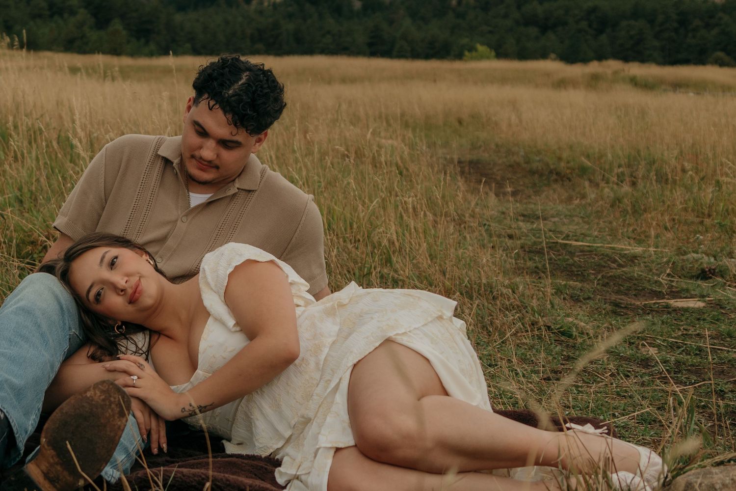 Woman rests head on mans lap while relaxing together in golden grass field during engagement photos.