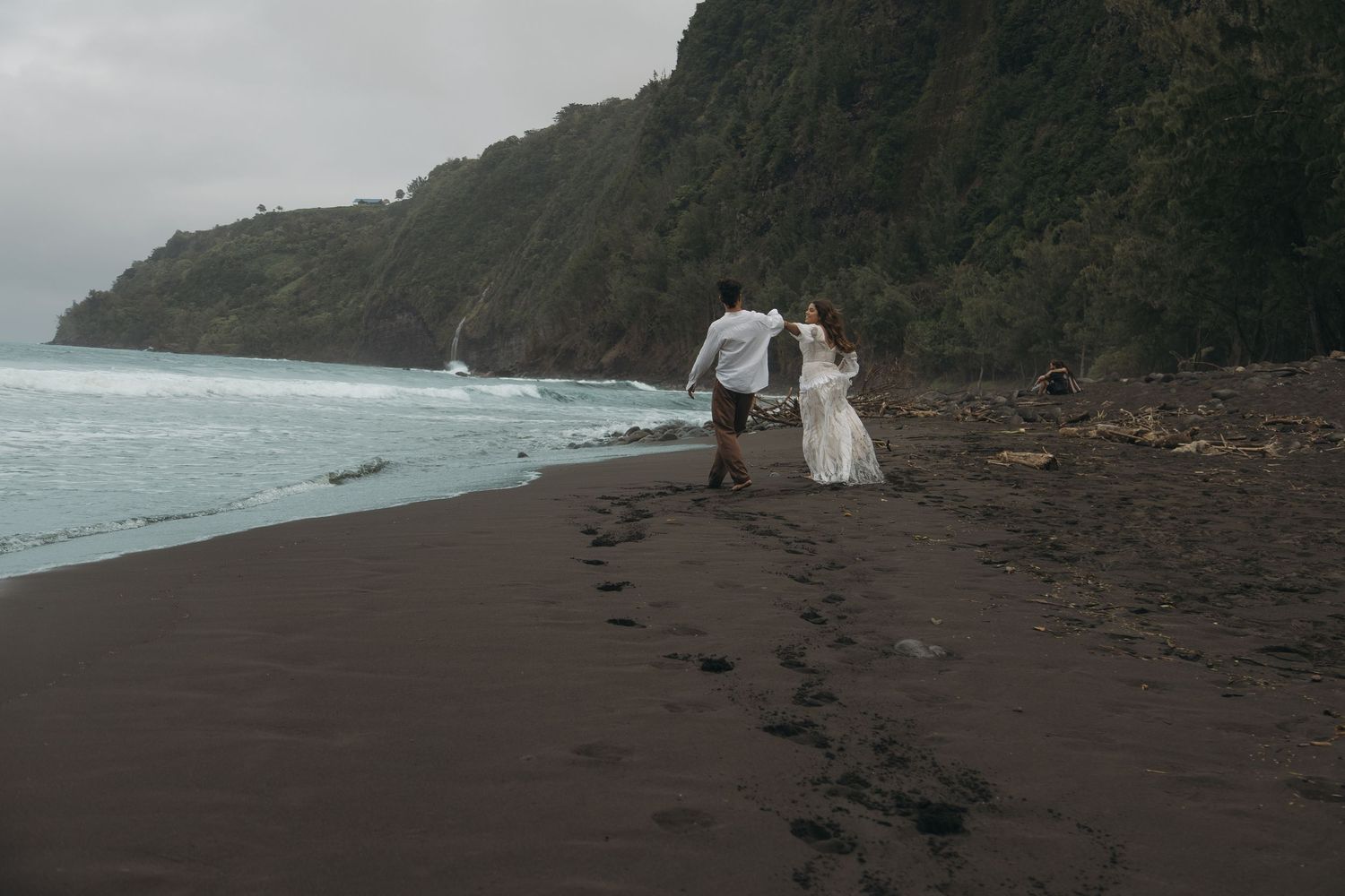 Couple in wedding attire walking hand in hand on a black sand beach with lush green cliffs and ocean waves.