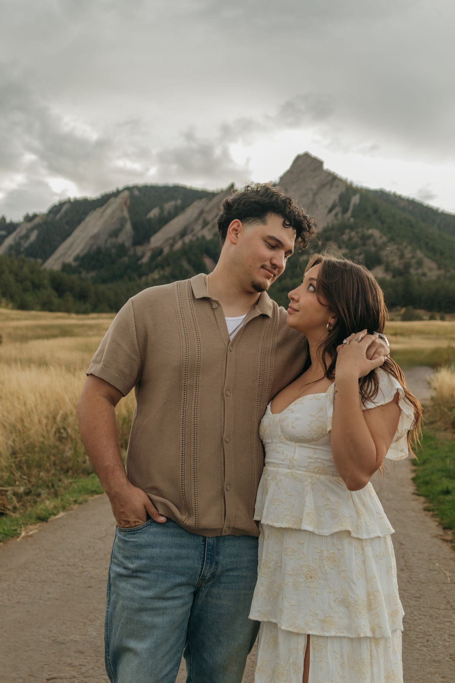 A couple walks along a path gazing at each other with the dramatic Flatirons mountains behind them.