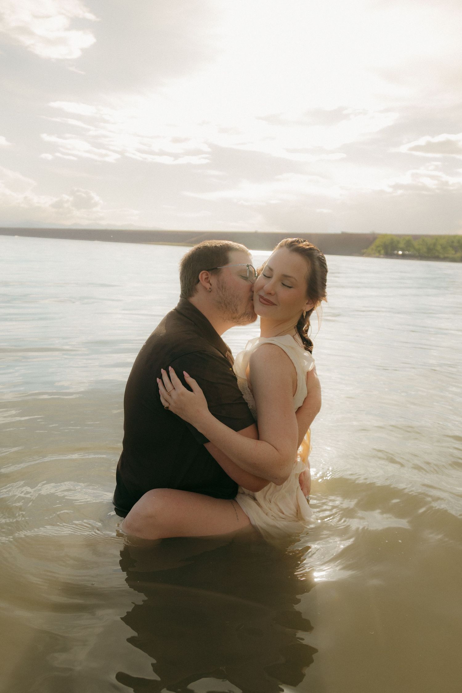 Couple embracing romantically in shallow water at sunset, woman smiling as man kisses her cheek.