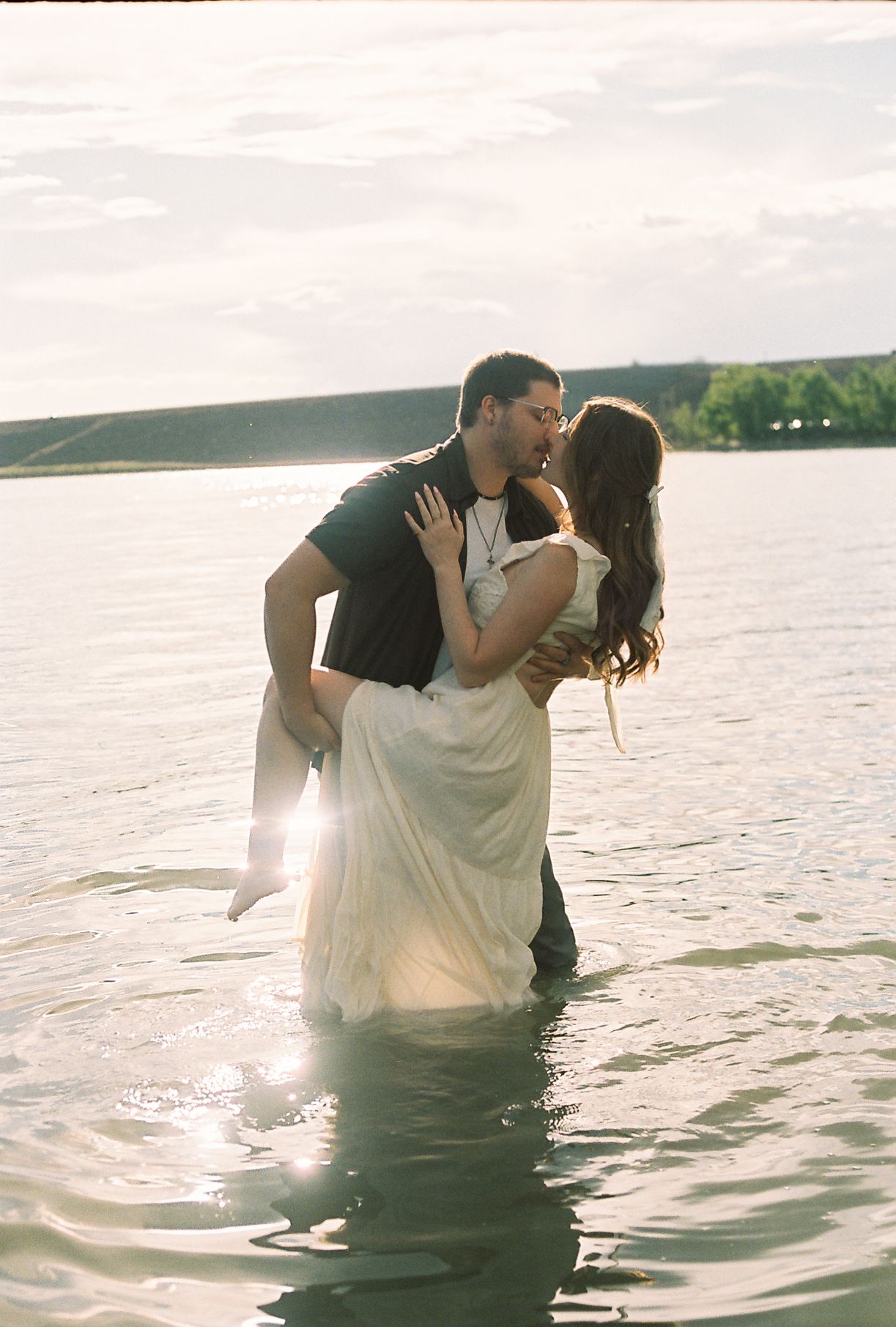 Groom lifting bride in white wedding dress while kissing in lake water with golden sunset light behind them.