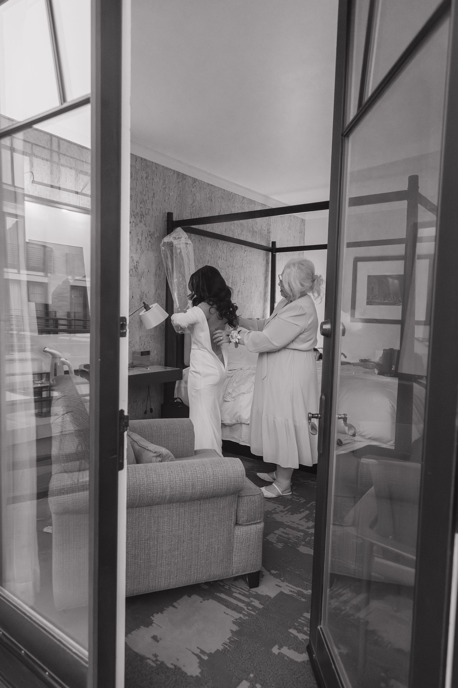 Black and white photo of a bride being helped into her wedding dress through French doors.
