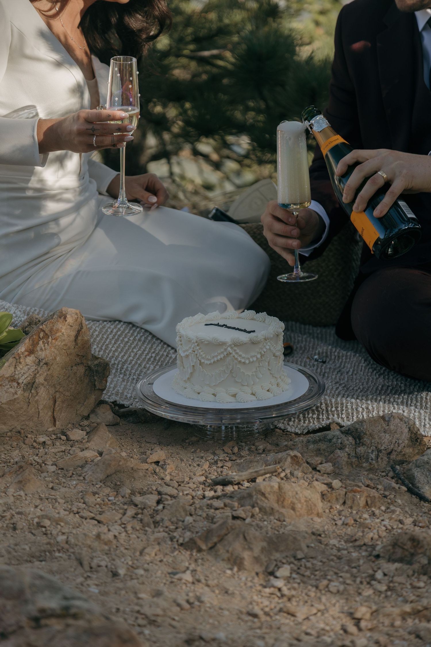 Bride and groom sitting outdoors on rocks, pouring champagne beside a small white wedding cake.