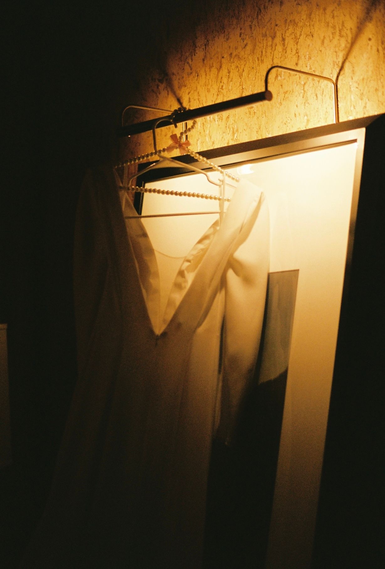 White wedding dress and veil hanging on a hanger under warm golden light in a dimly lit hotel room.