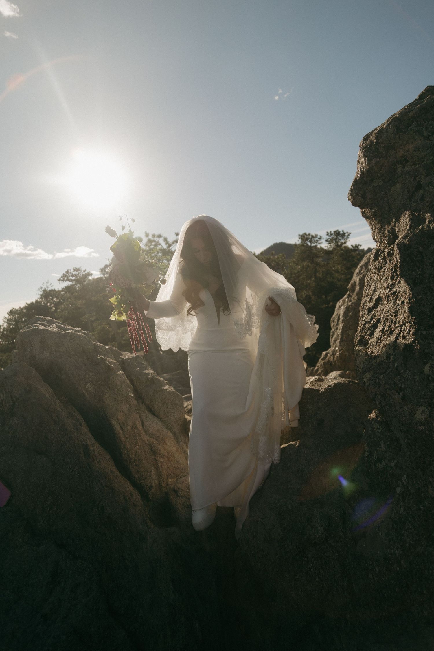Bride in white gown and veil standing among rocks with sun flare creating a dreamy backlit effect.
